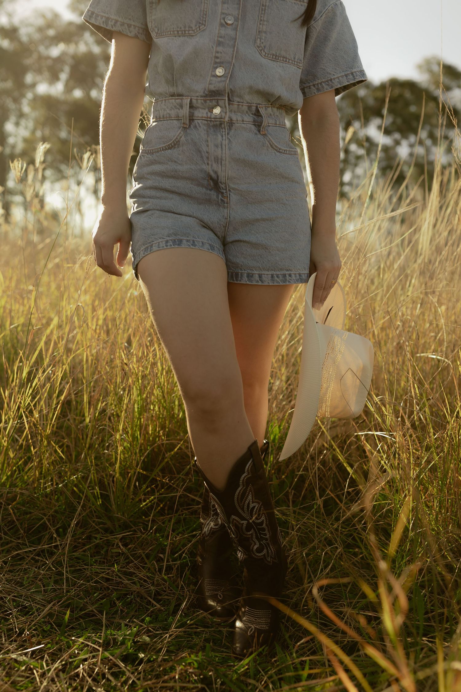 Woman in denim shorts, cowboy boots, holding straw hat, standing in golden grass field at sunset.