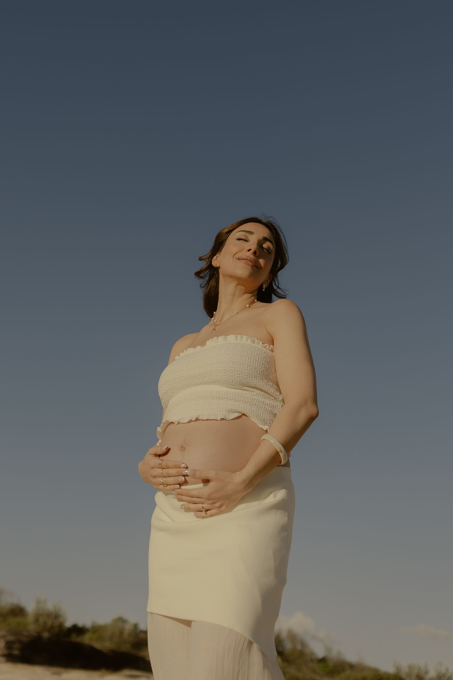 Pregnant woman in white strapless top and skirt cradling her baby bump outdoors under a clear blue sky.