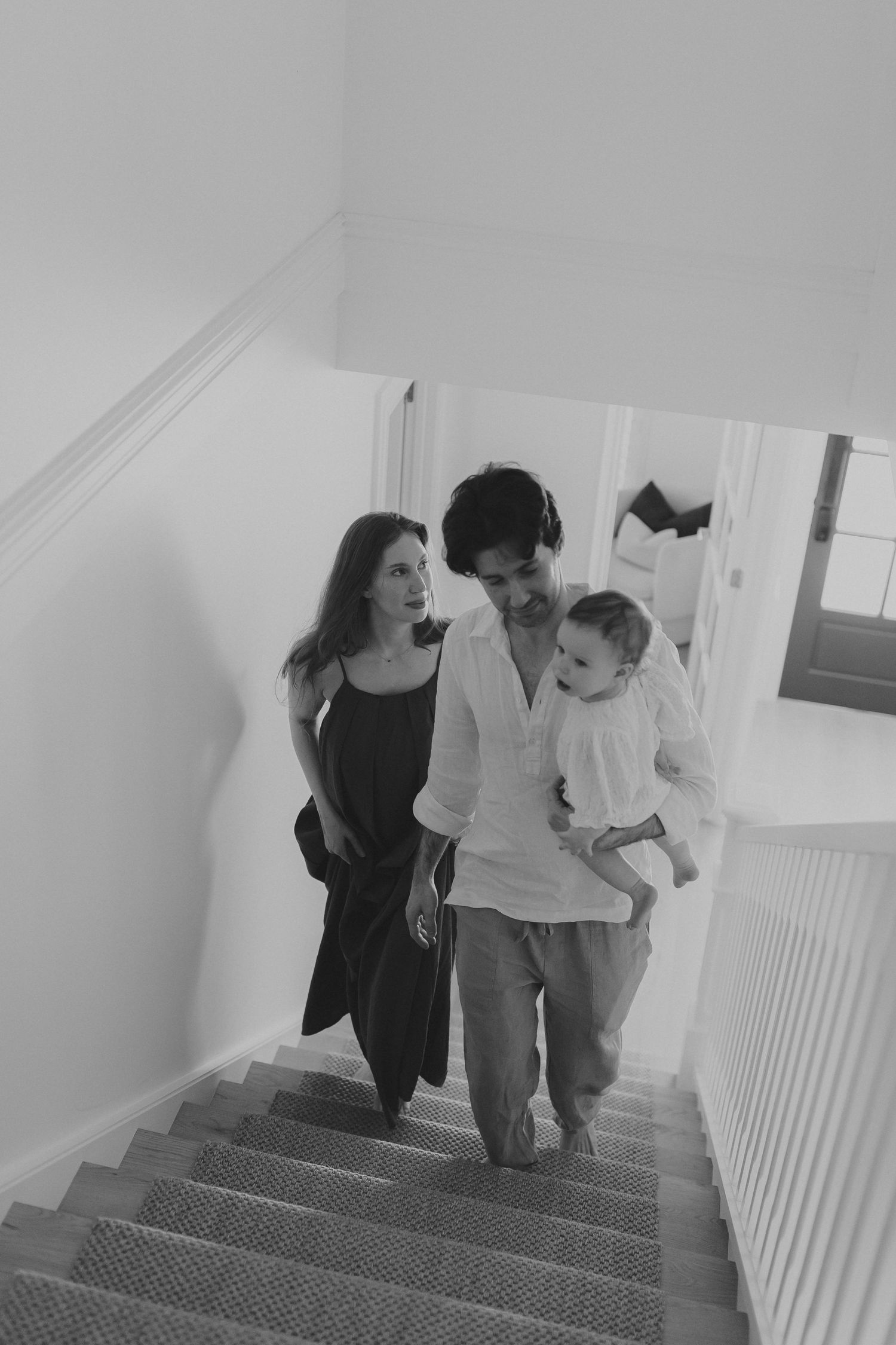 Black and white photo of a young family walking up stairs, man carrying baby while woman looks on smiling.
