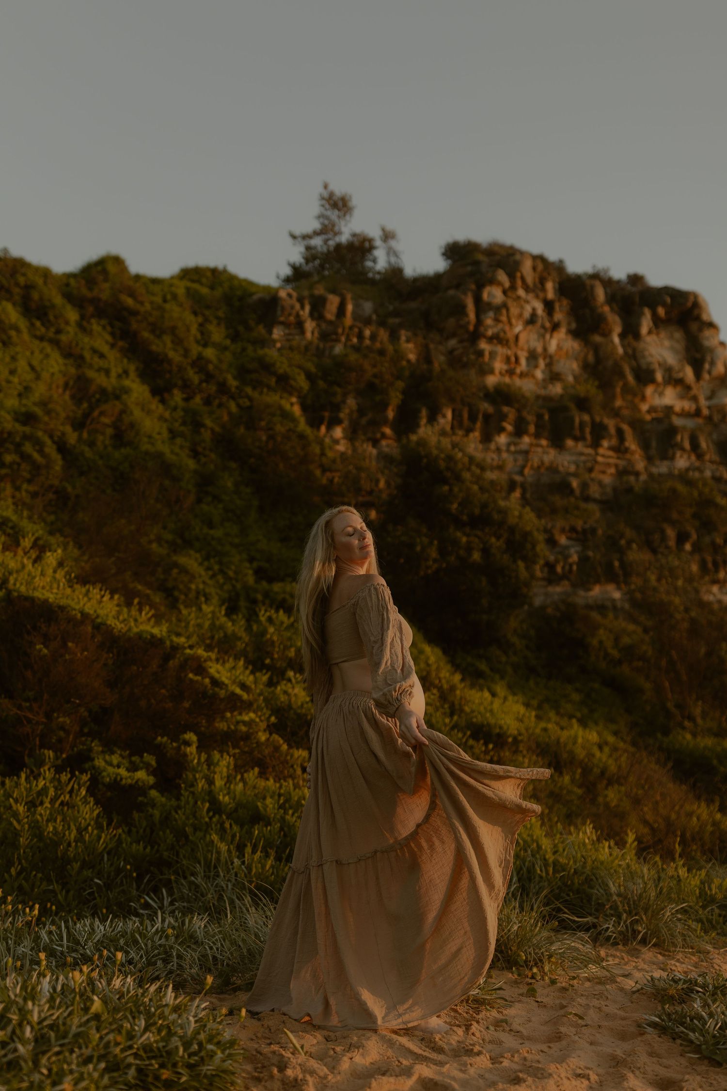 Pregnant woman in flowing beige dress standing on sandy path with rocky cliffs and lush greenery at golden hour.