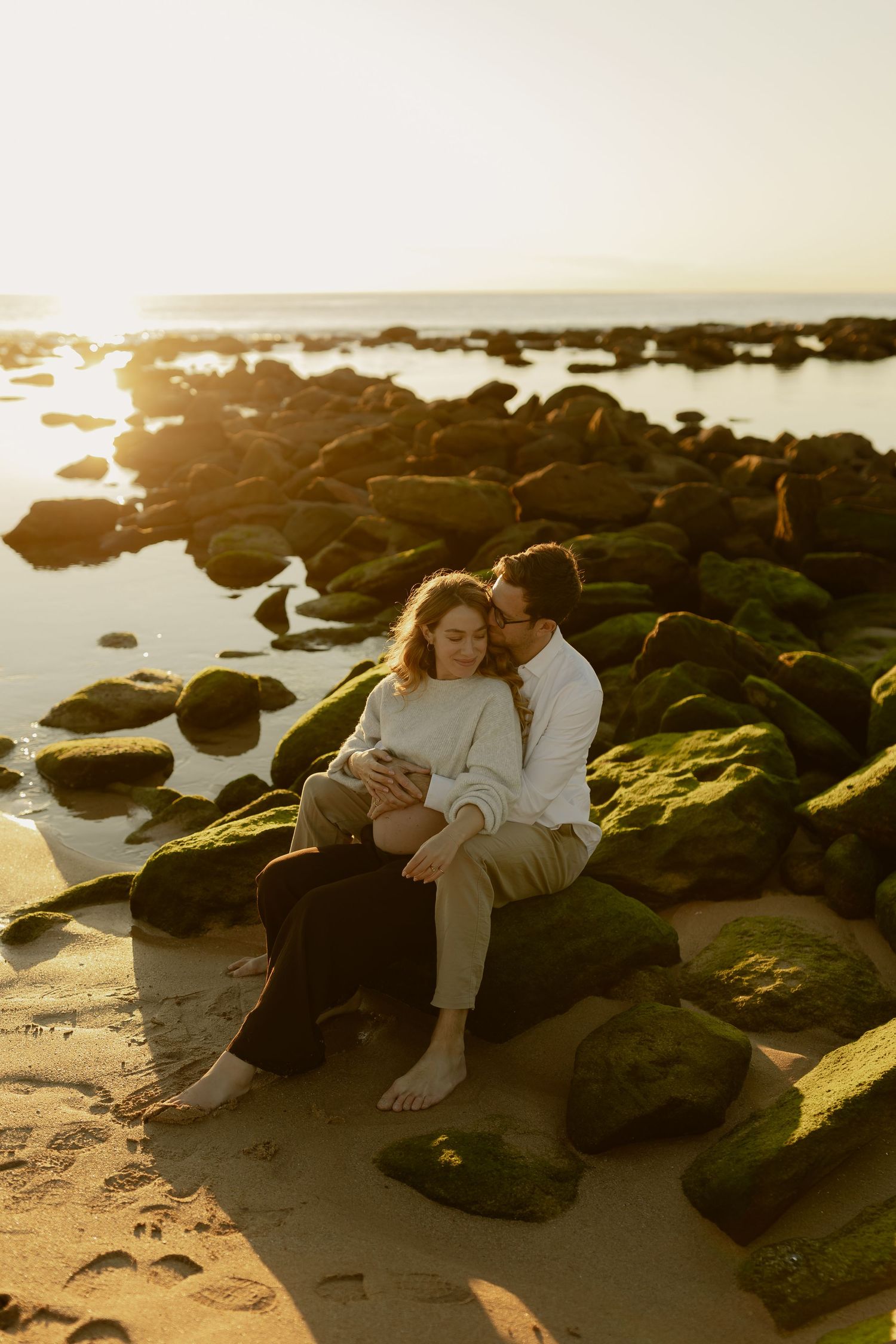 Couple sitting together on moss-covered rocks at beach sharing tender moment at sunset.
