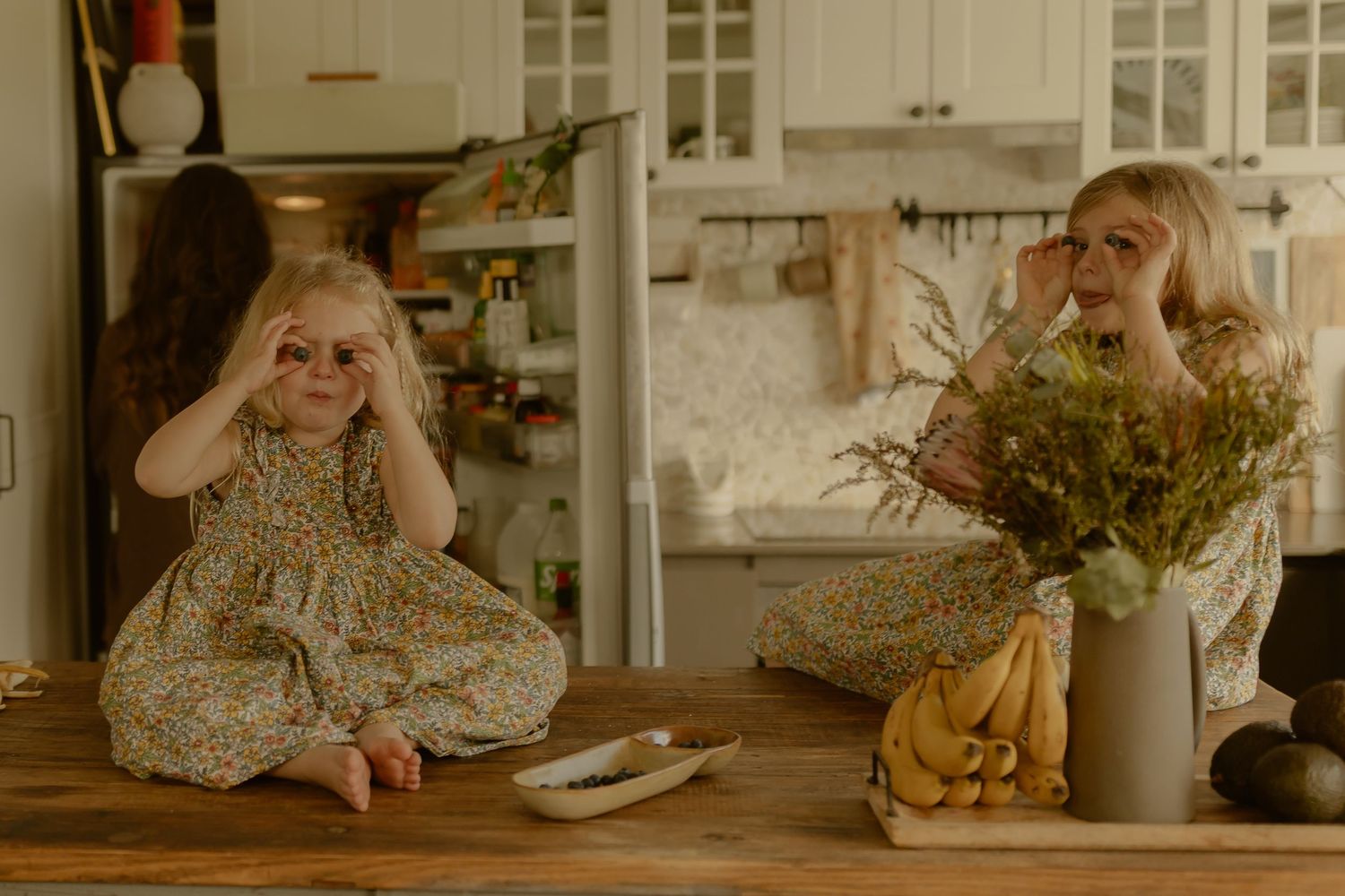 A woman and young girl in floral outfits sit on a kitchen counter making silly faces together.