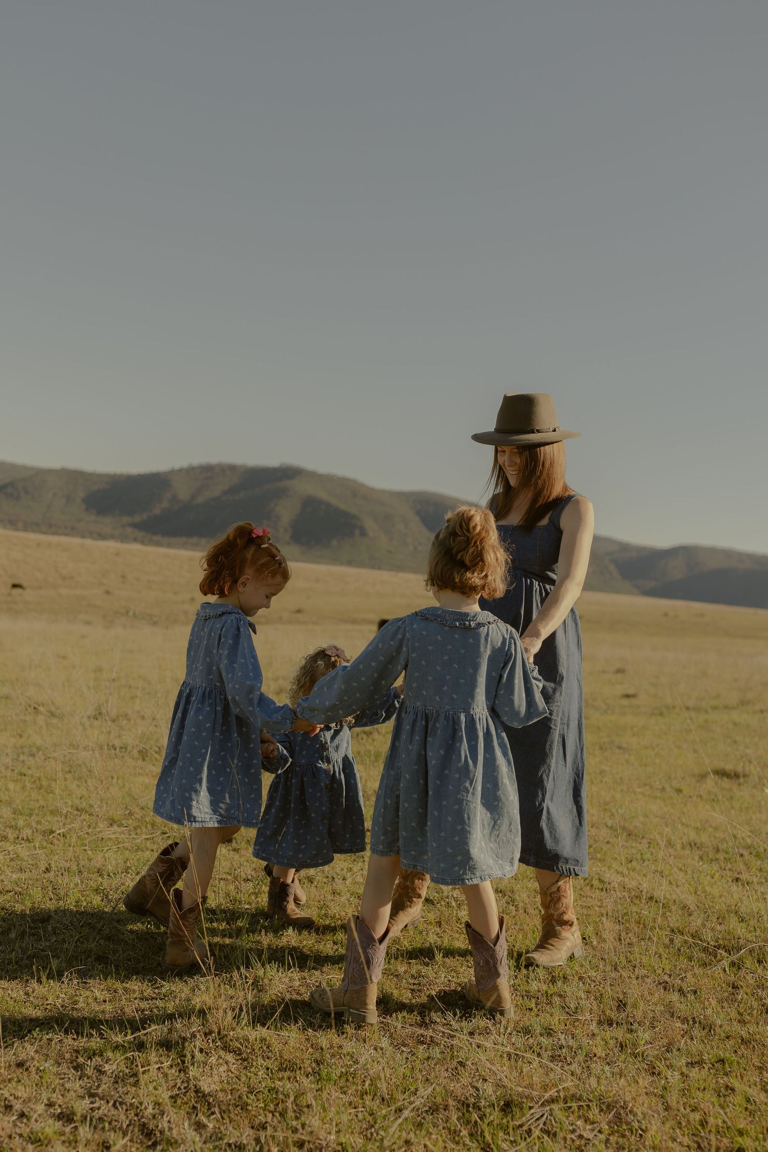 A woman in a hat and blue dress plays with two young girls in matching dresses on a grassy field with mountains.