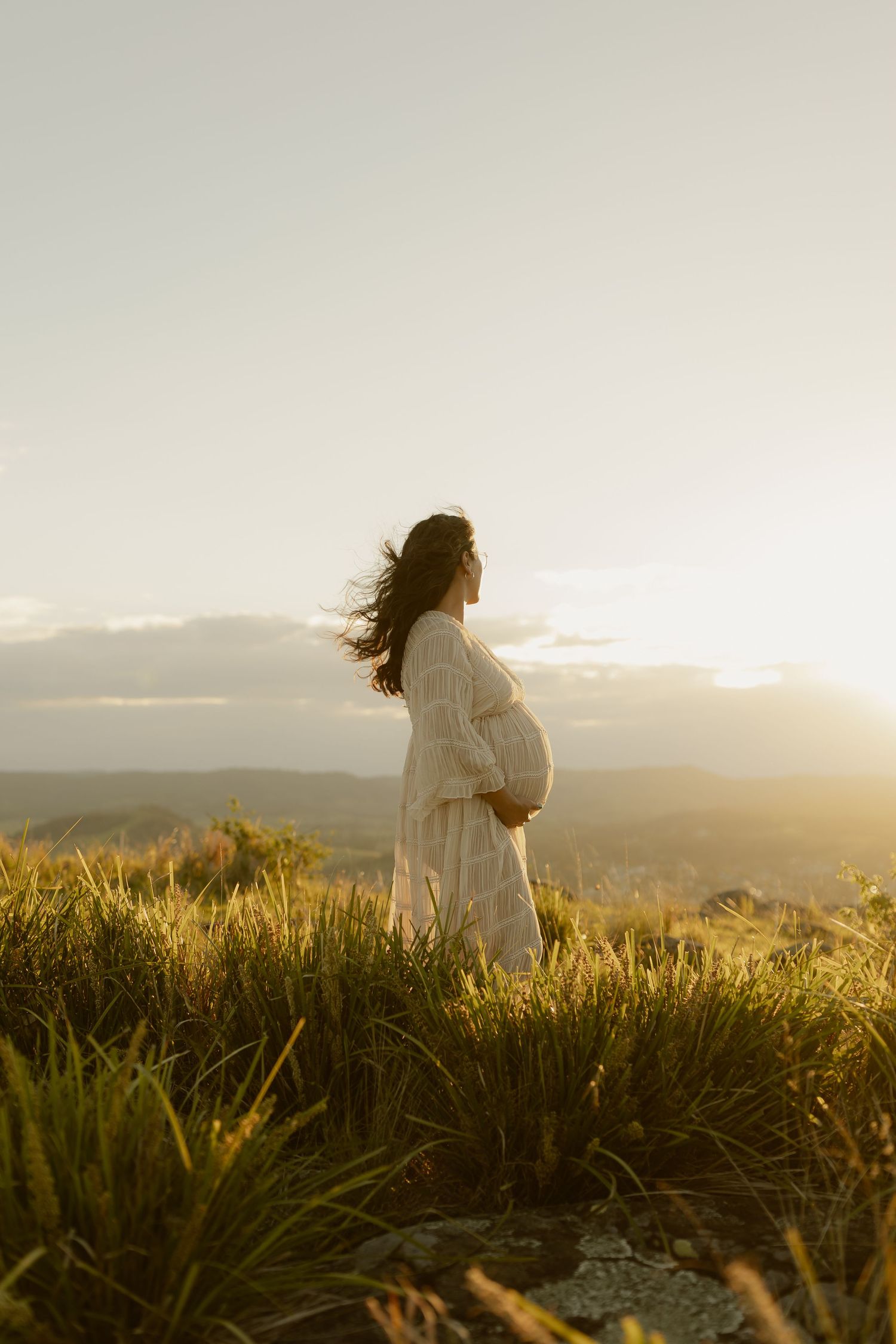 Pregnant woman in white dress cradling belly on grassy hilltop at golden sunset.