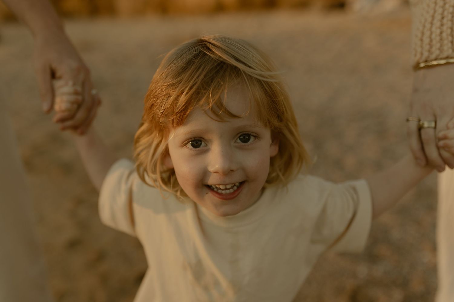 Happy red-haired child smiling while holding hands with adults outdoors in warm golden light.