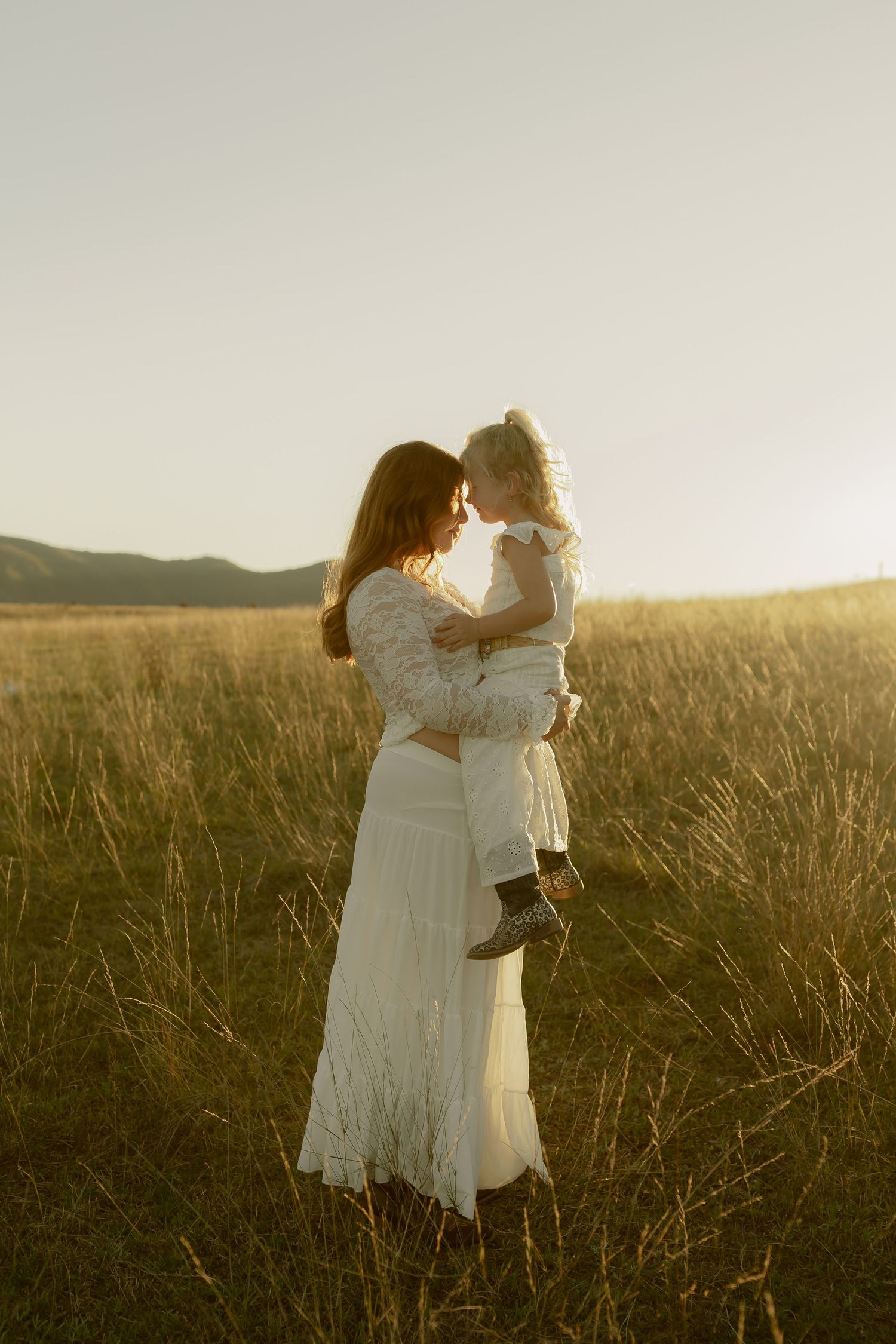 A mother in white holds her young child tenderly in a sunlit golden meadow with hills behind.