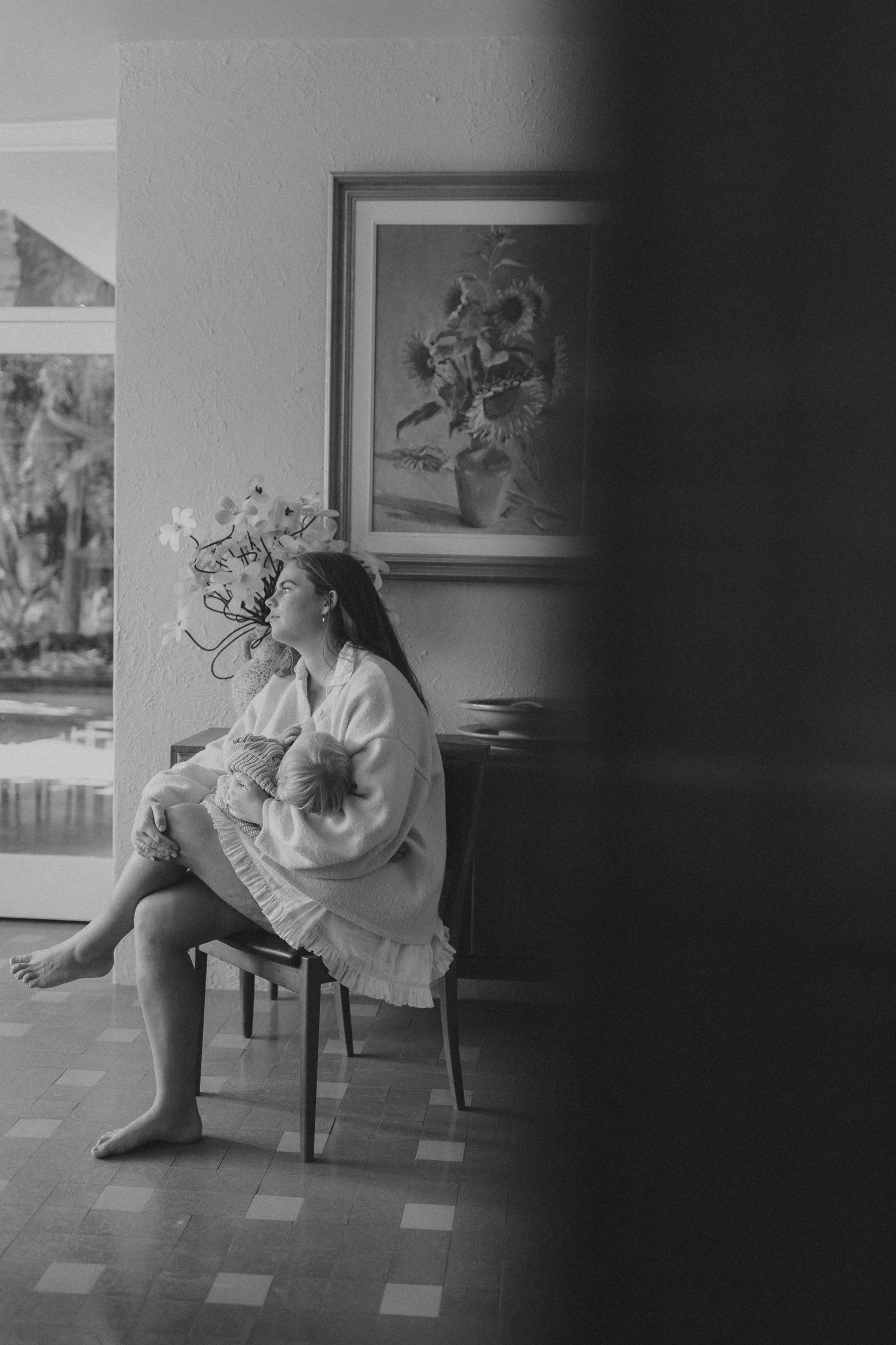 Black and white photo of a mother in a white robe sitting on a chair holding her newborn baby near a window.