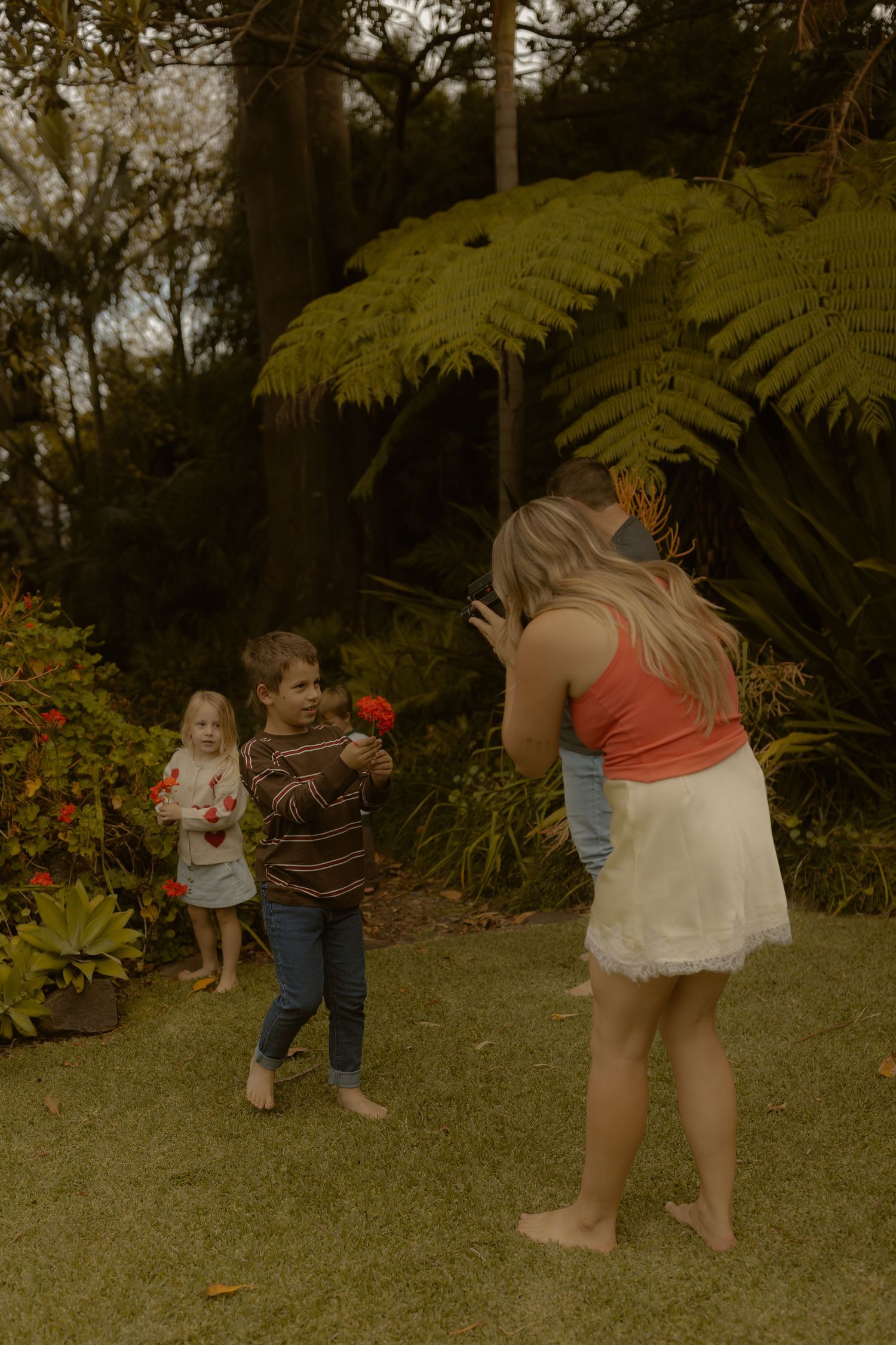 Children playing in a lush garden, with a boy holding a red flower while a woman and toddler watch nearby.