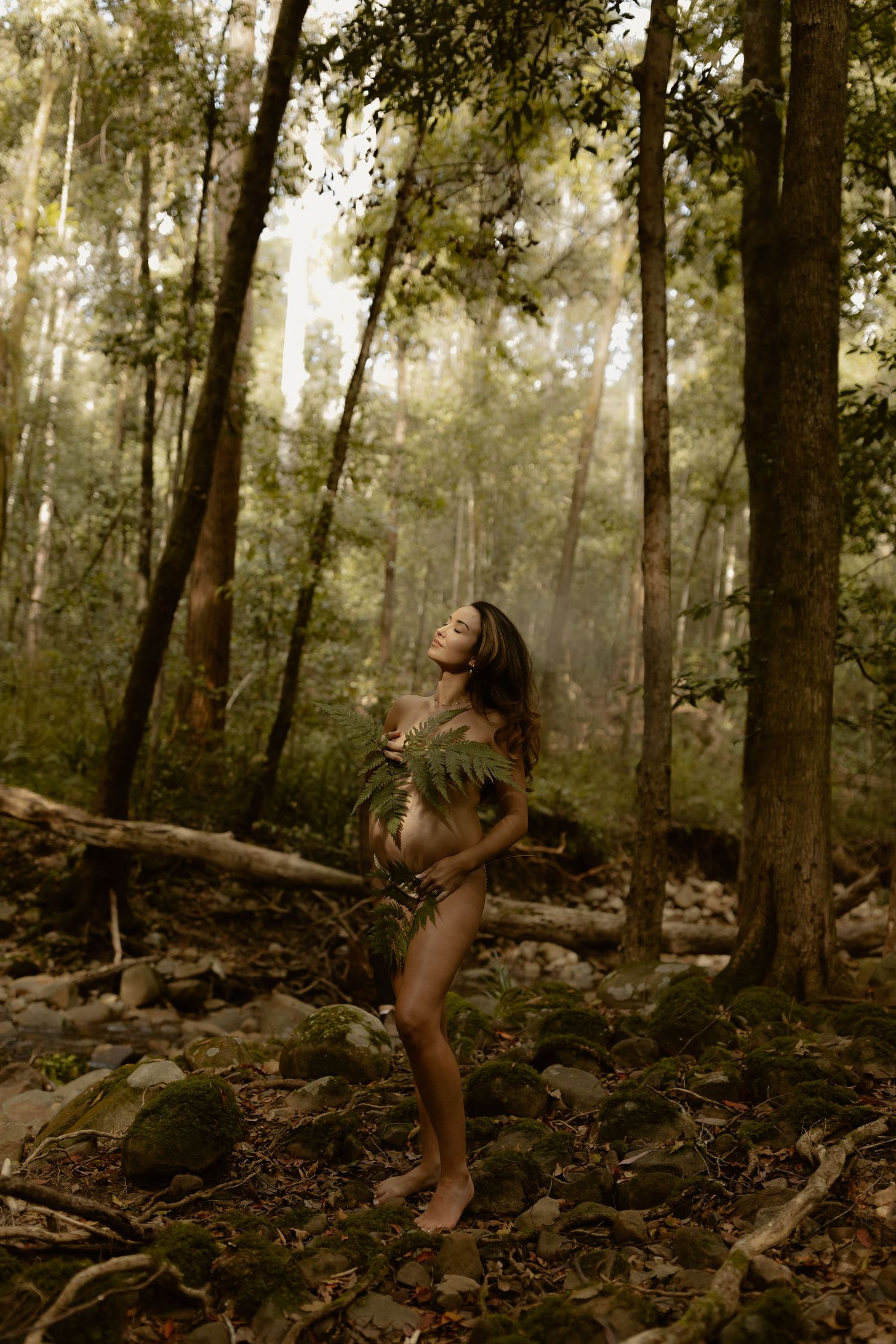 Pregnant woman poses gracefully in a misty forest, adorned with ferns, surrounded by tall trees and mossy rocks.