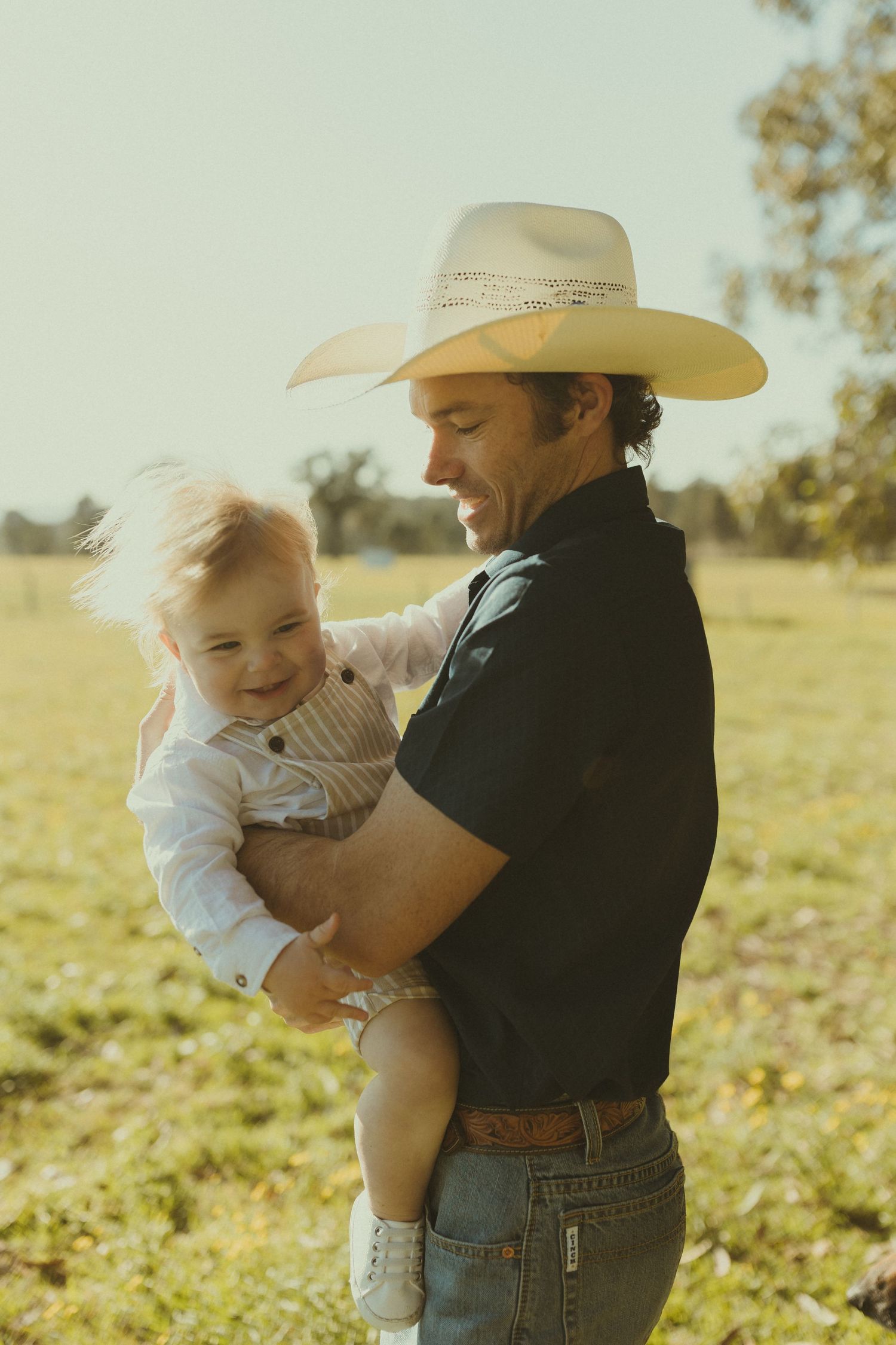 A cowboy in a straw hat holds a small child wearing striped overalls in a sunny field.