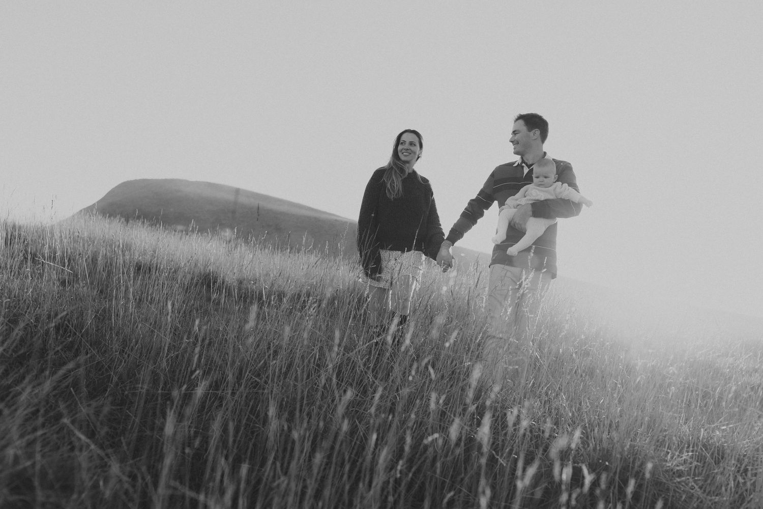 A black and white photo of a couple holding hands and carrying their newborn in a grassy field.