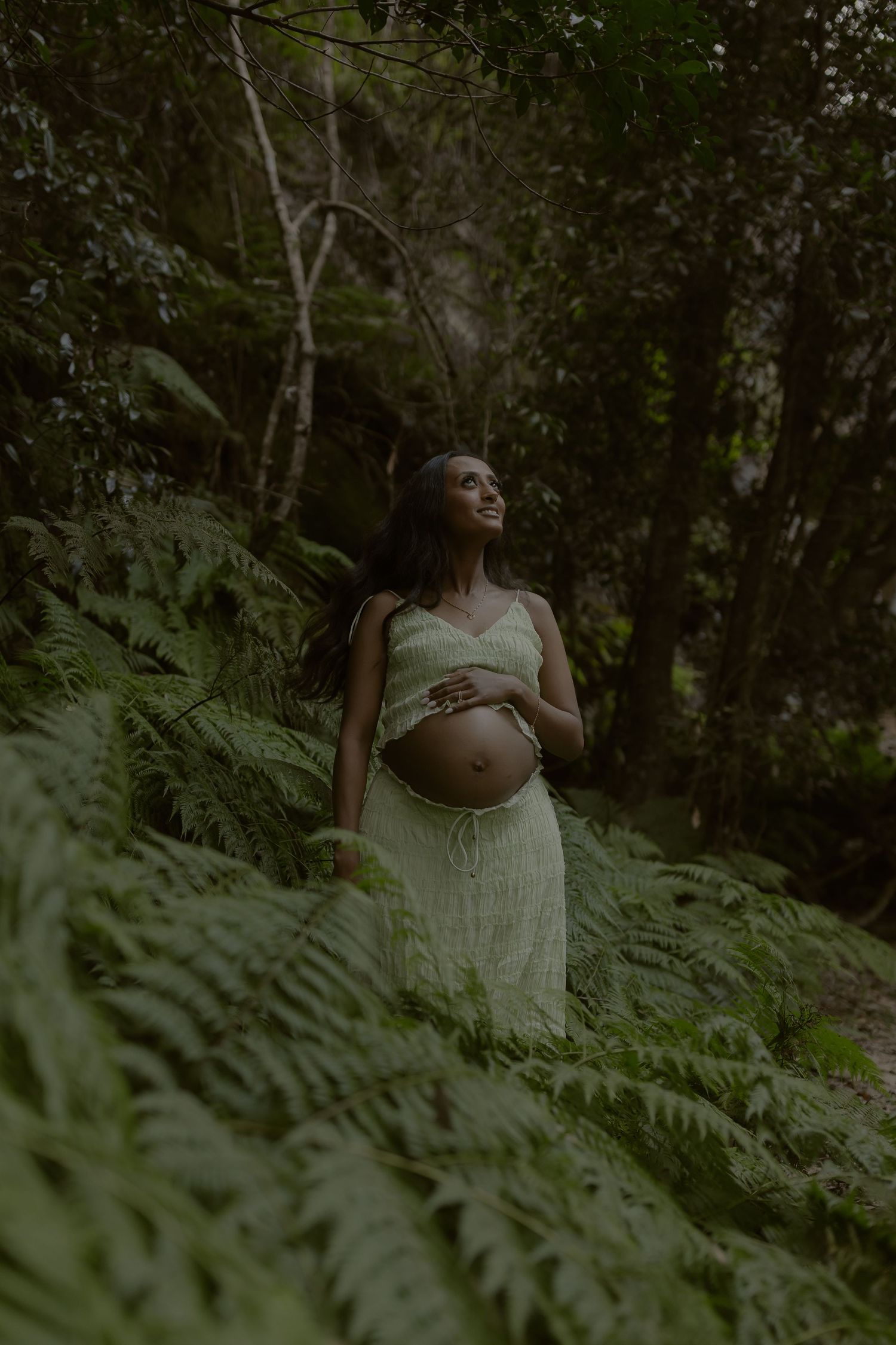 Pregnant woman in green two-piece dress cradling baby bump among lush forest ferns.
