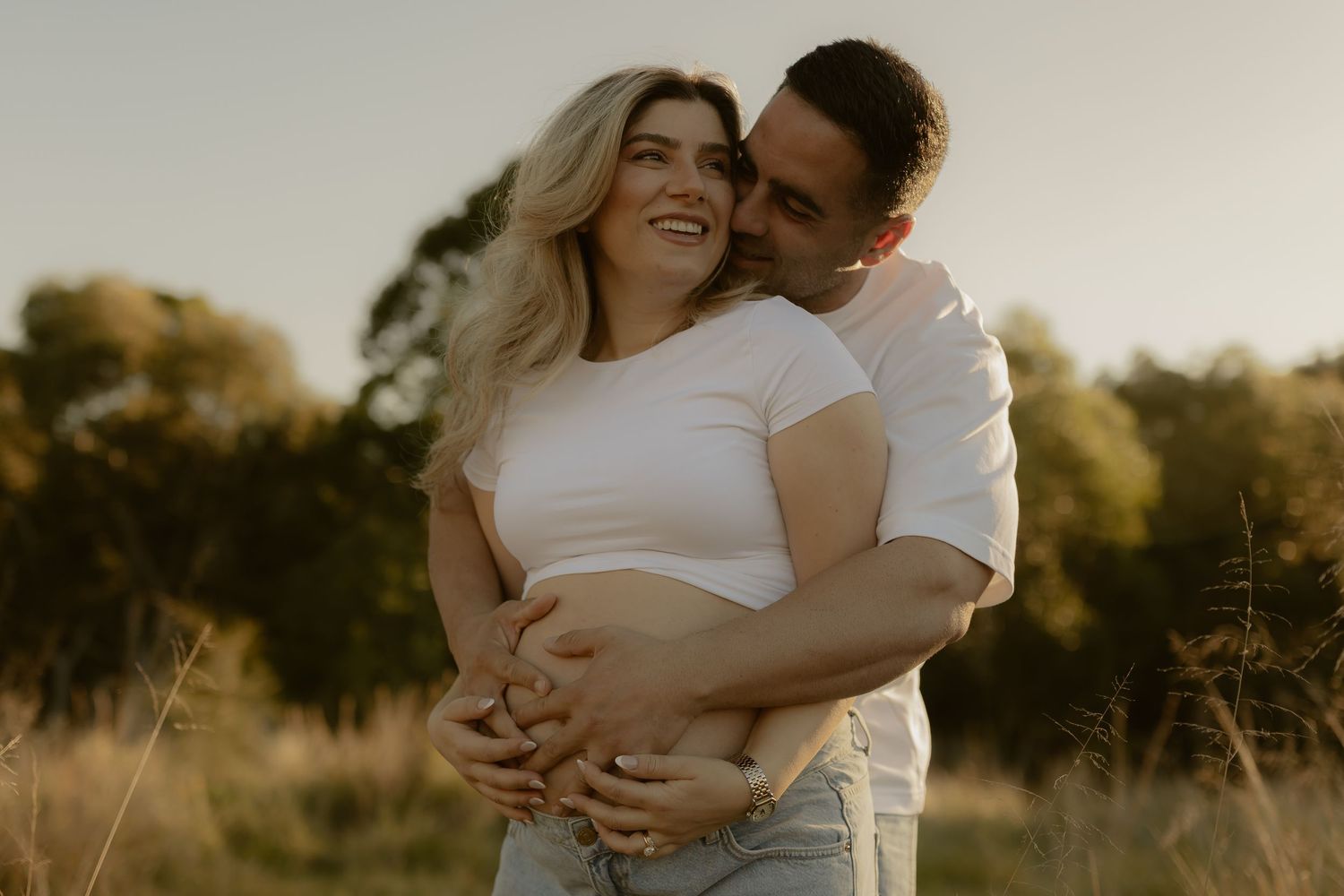 Happy couple embracing outdoors at golden hour, man hugging pregnant woman from behind in a sunny field.