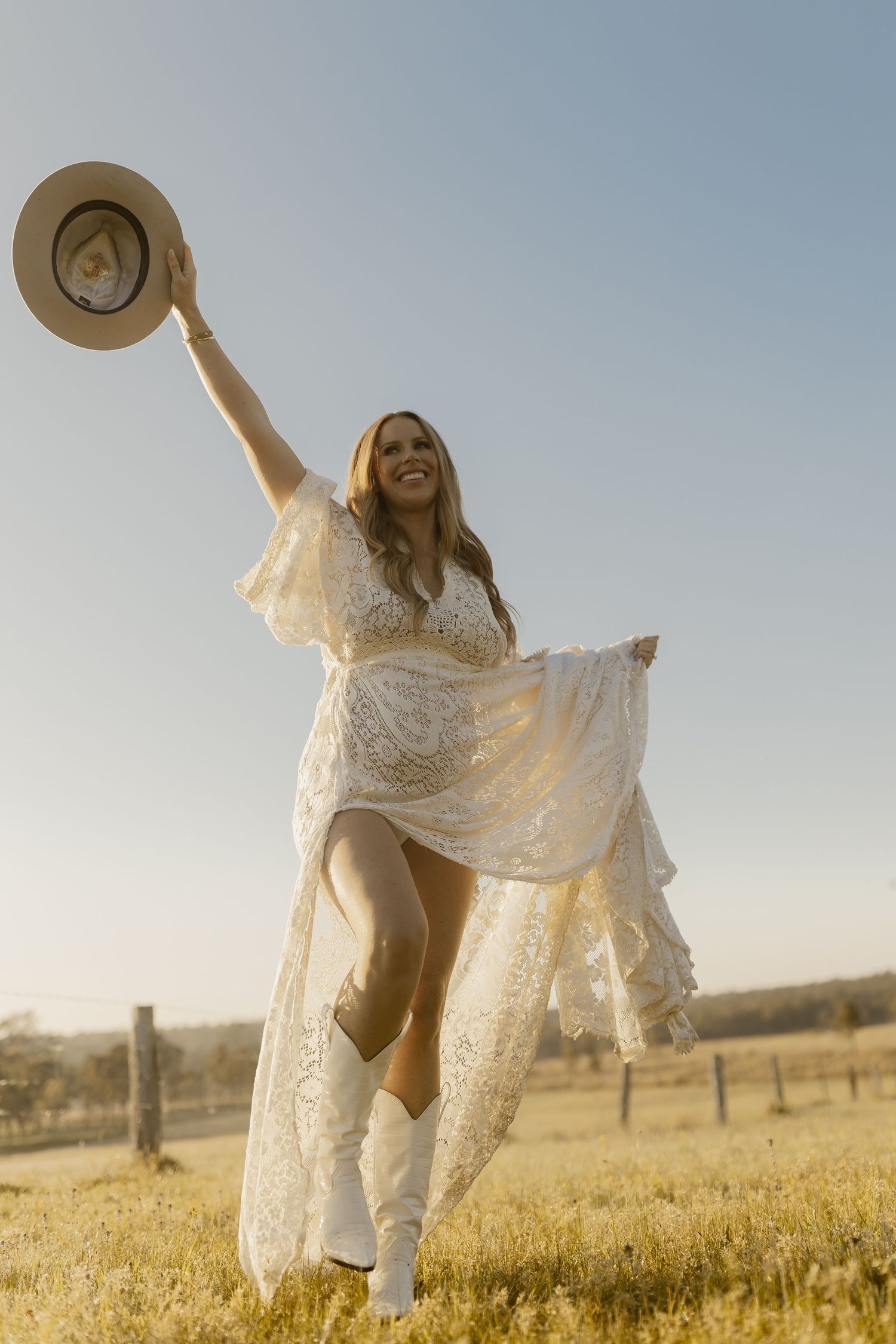 A joyful woman in a white lace dress and cowboy boots raises a hat in a sunny field.