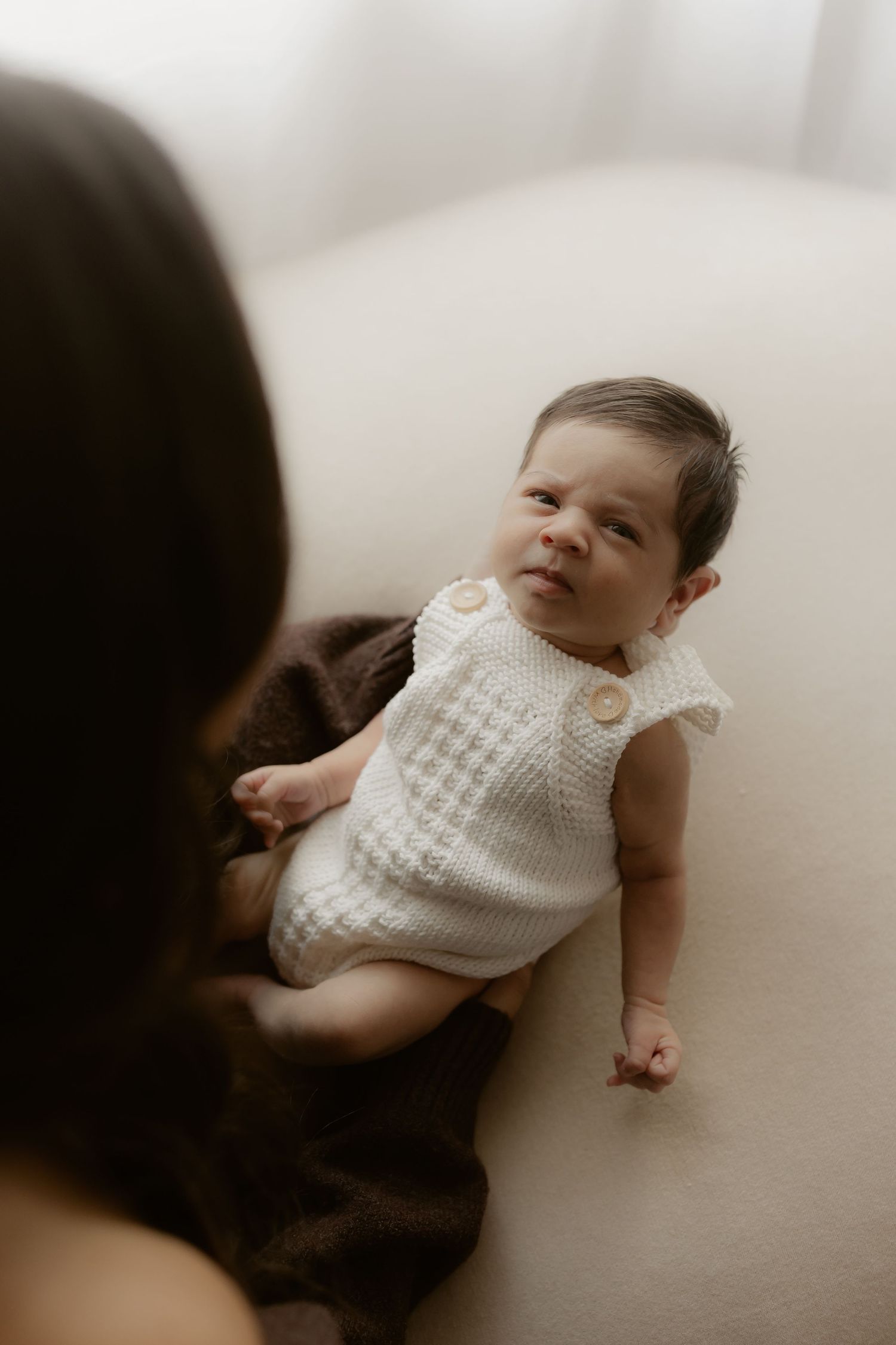 Newborn baby in white knit romper lying on cream surface, looking up at mother during professional newborn photo session.