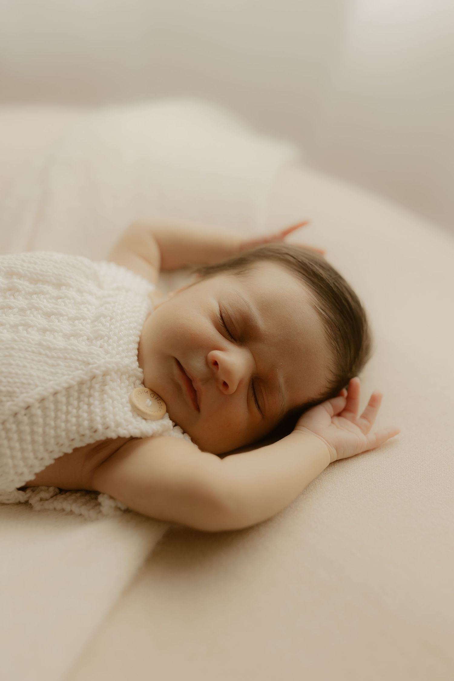 Peaceful newborn baby sleeping on a soft white surface wearing a cozy cream knit romper.