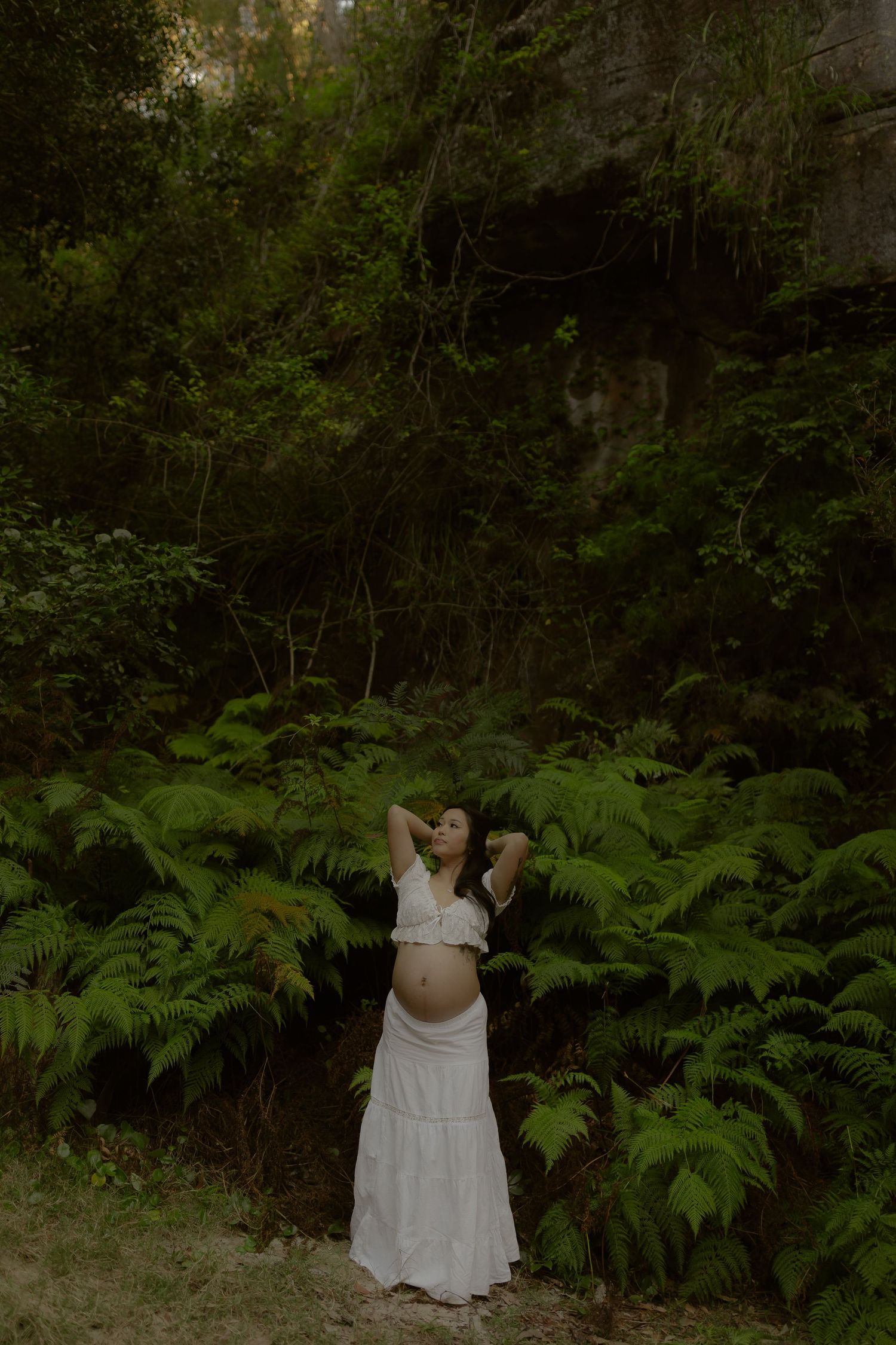 Pregnant woman in white crop top and skirt standing among lush ferns in a forest, looking up gracefully.