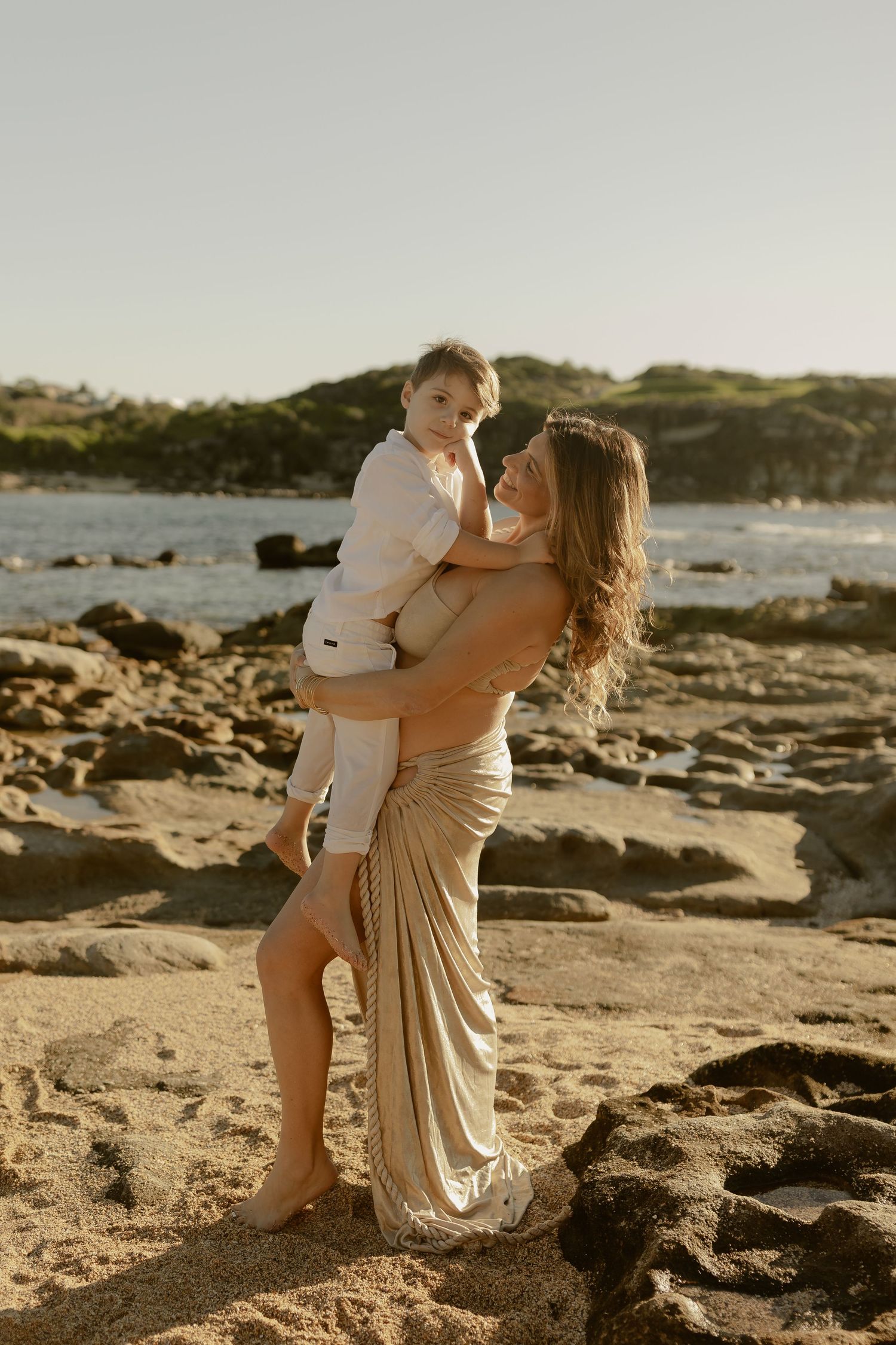 A woman in a gold dress holds a young boy on a rocky beach at sunset.