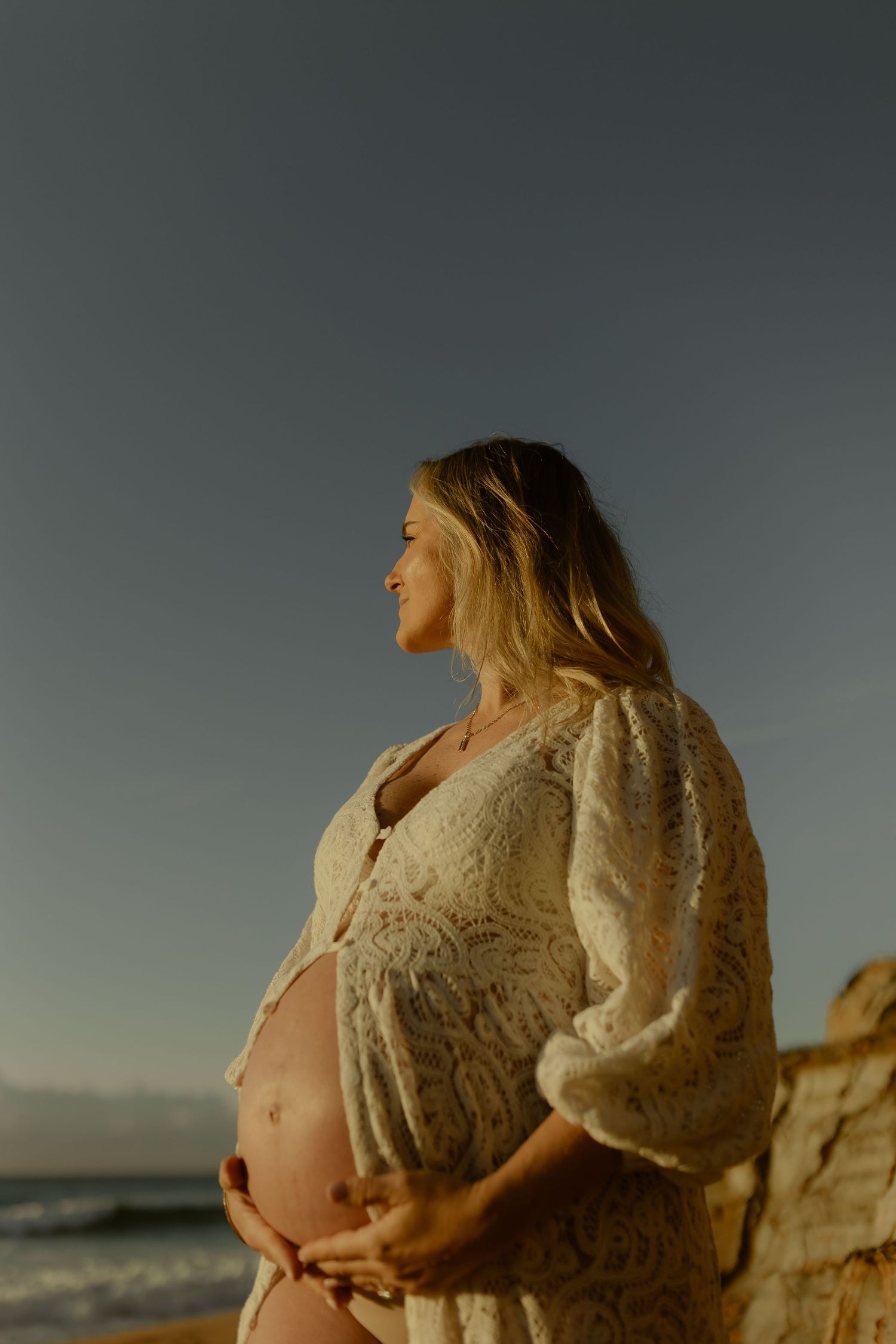 Smiling pregnant woman in white lace dress cradling her baby bump on a beach at golden hour sunset.