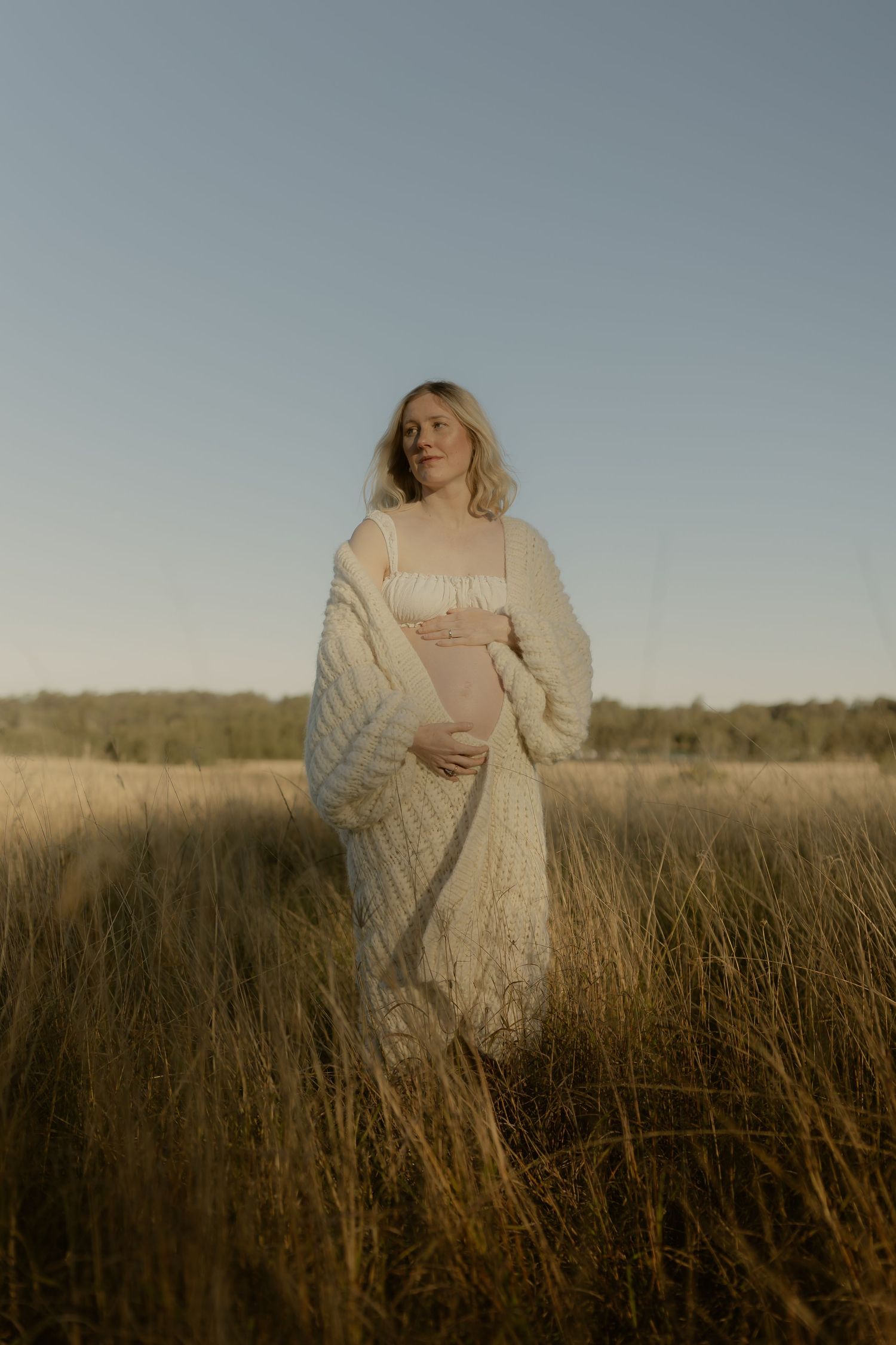 Pregnant woman in cream cardigan standing in golden grass field at sunset, cradling her baby bump.