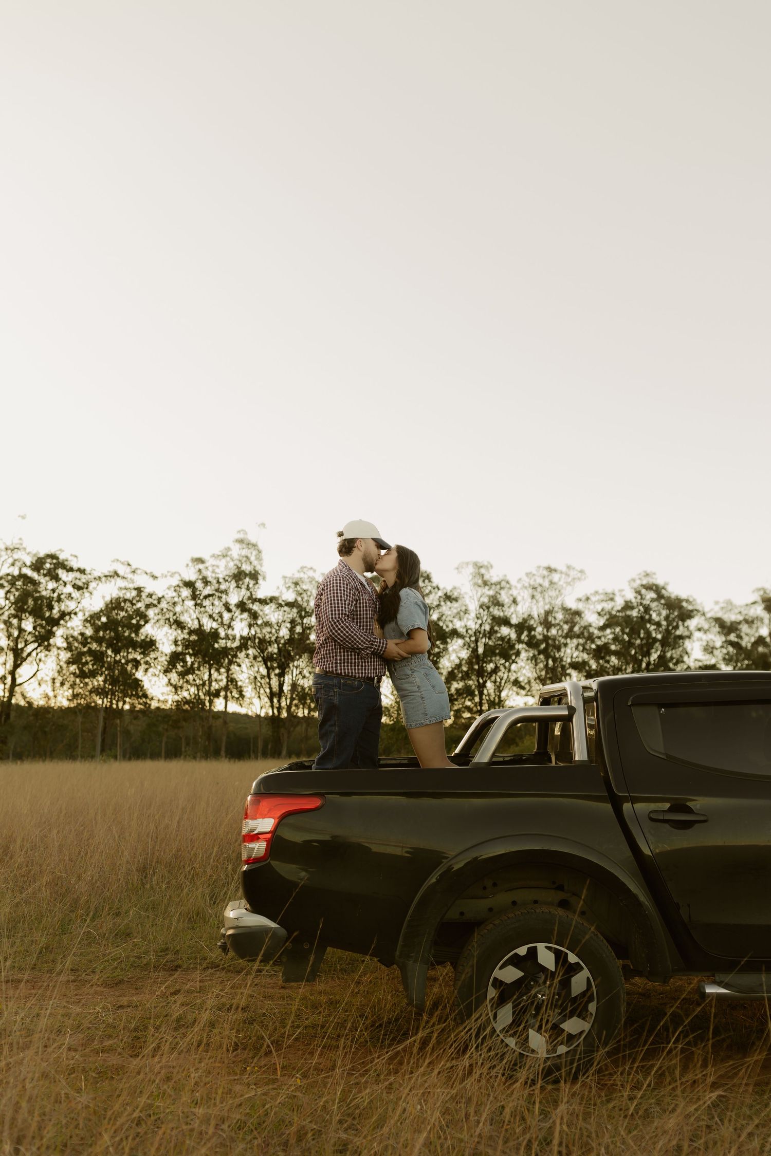 Couple kissing while standing in truck bed surrounded by tall grass at sunset.