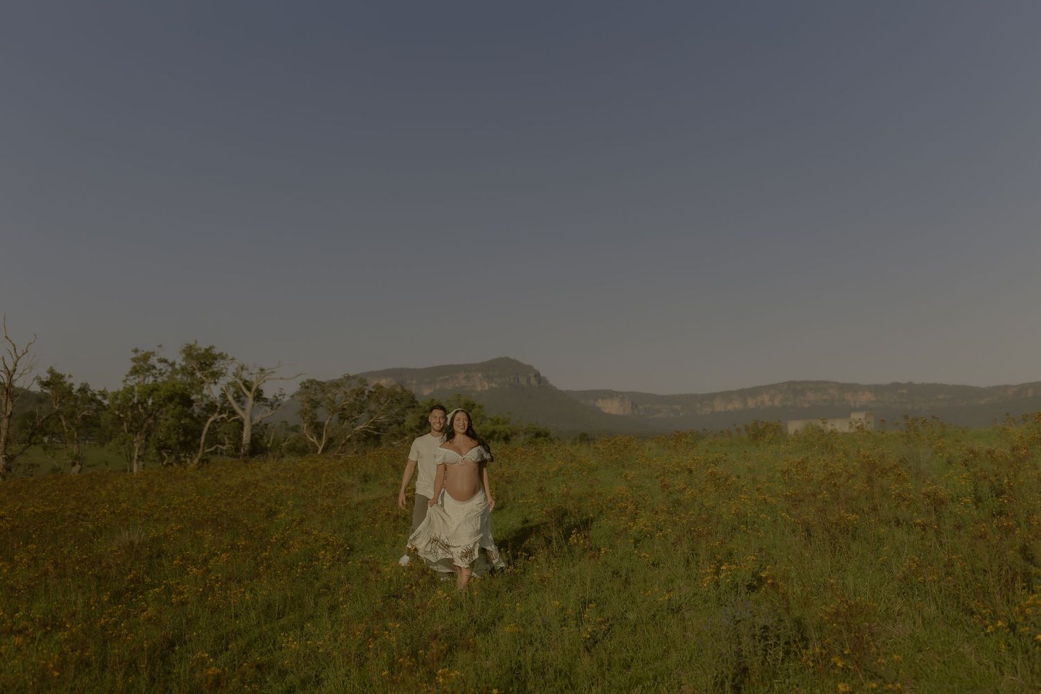 Couple in white outfits embrace in a sunny meadow with mountains in the background during golden hour.
