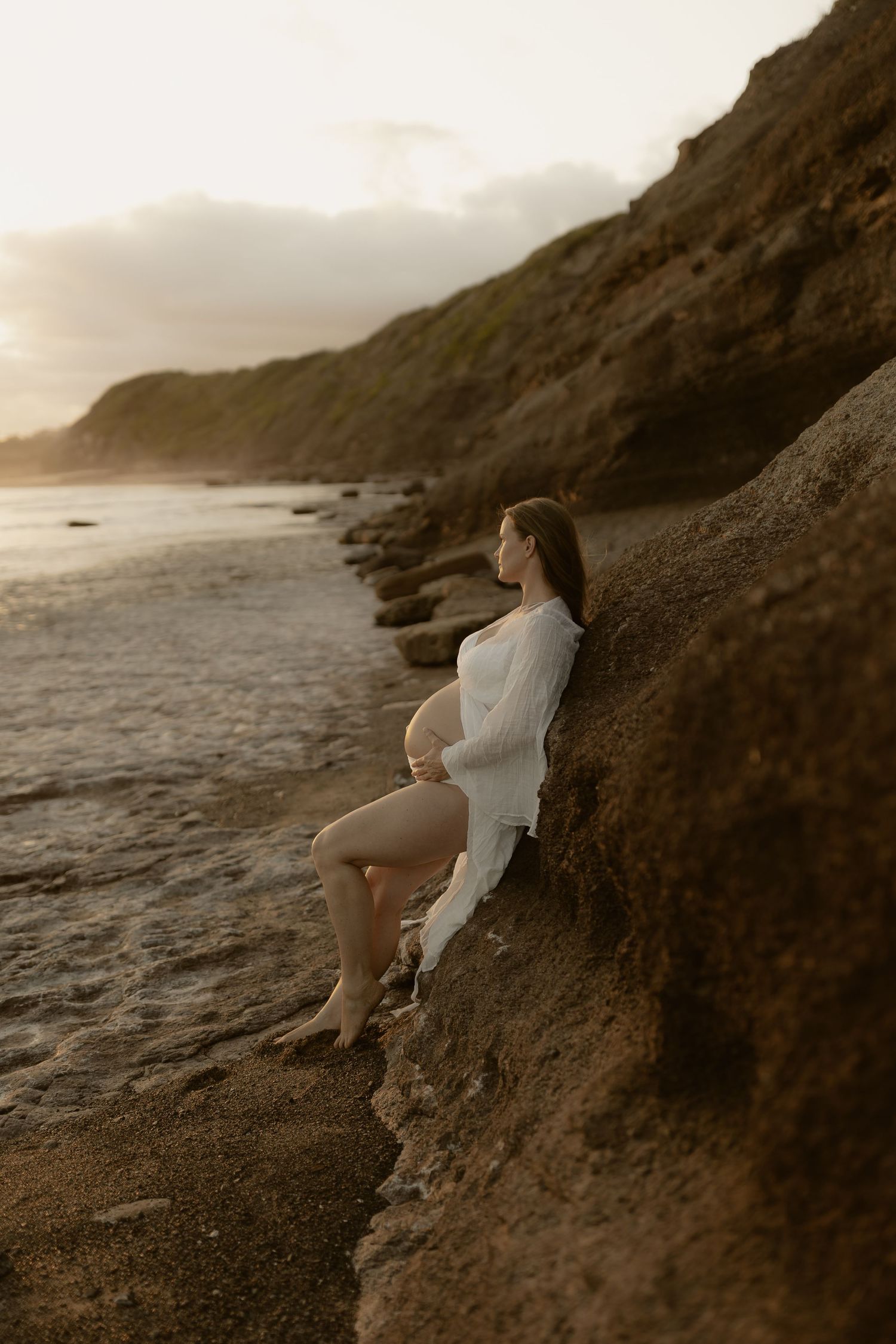 Person in flowing white dress perches on rugged coastal rocks as golden light illuminates the dramatic seascape.
