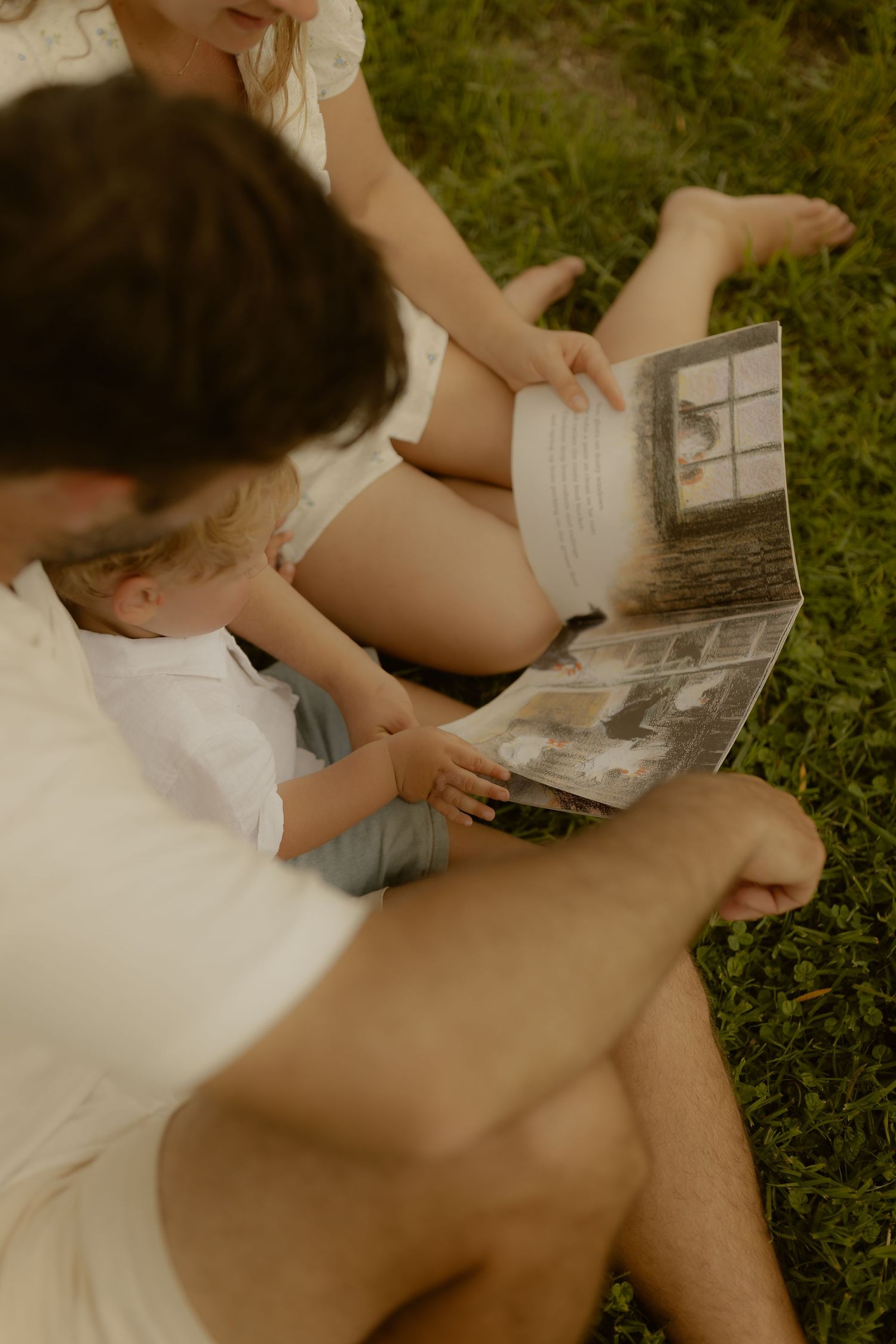 Children in white clothing read a book together while relaxing on grass outdoors.