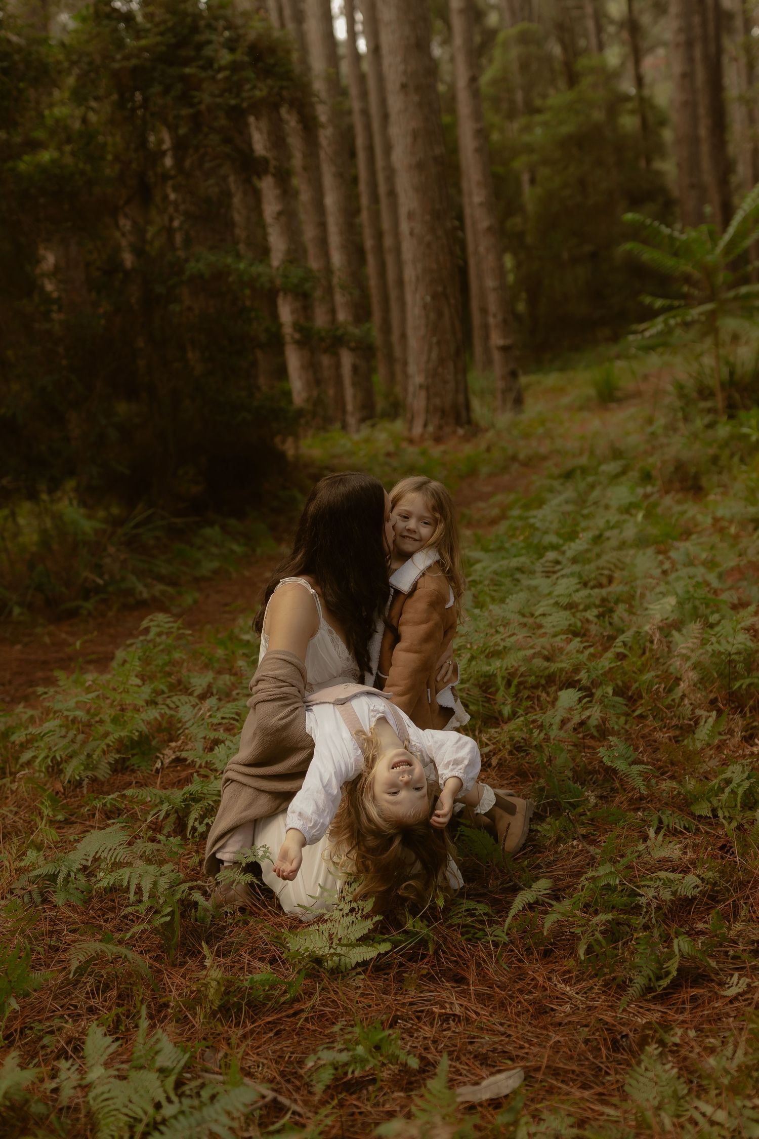 Mother playfully holding child upside down while another child watches in pine forest.