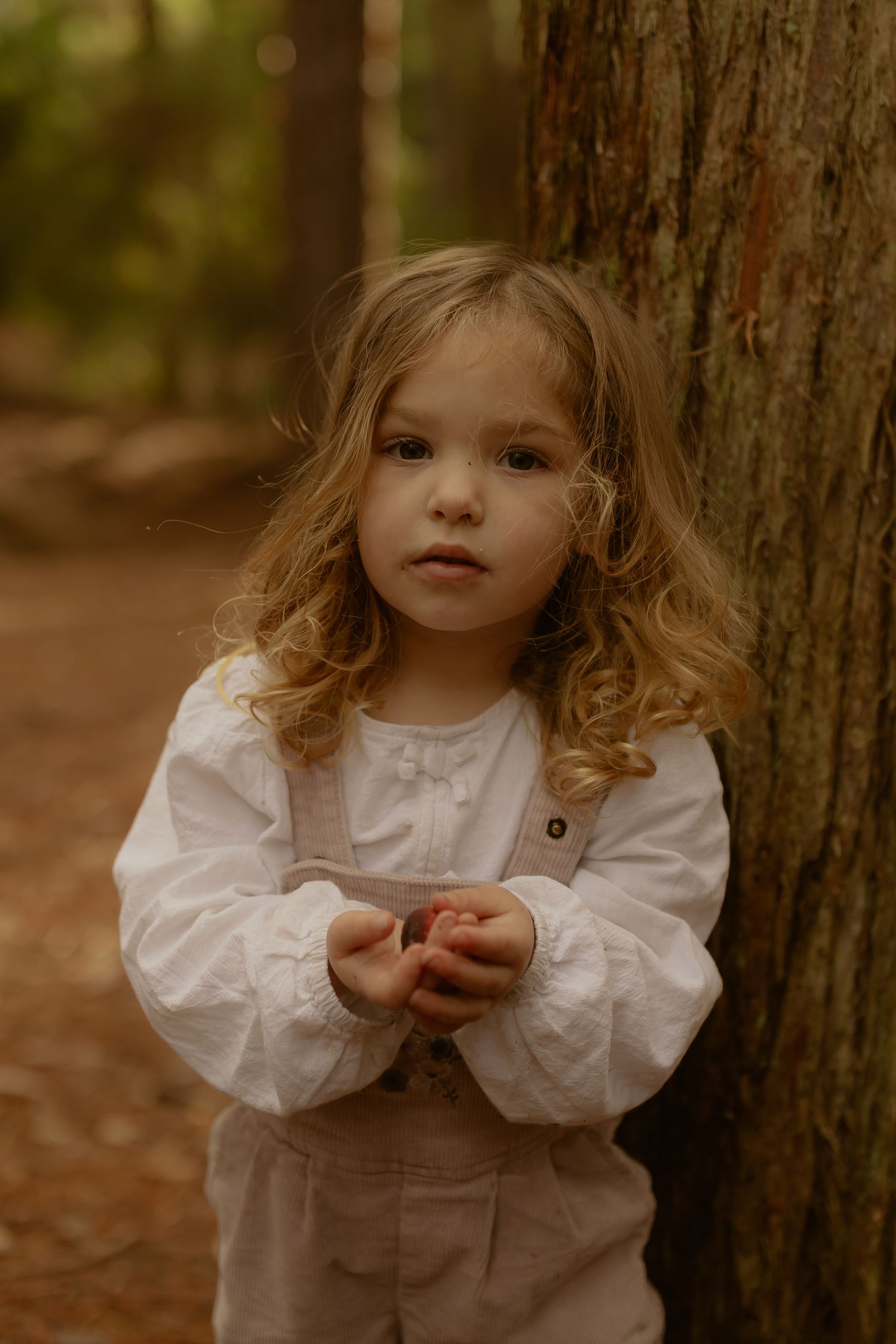 A young curly-haired girl in a white blouse and linen overalls leans against a tree trunk in a forest.