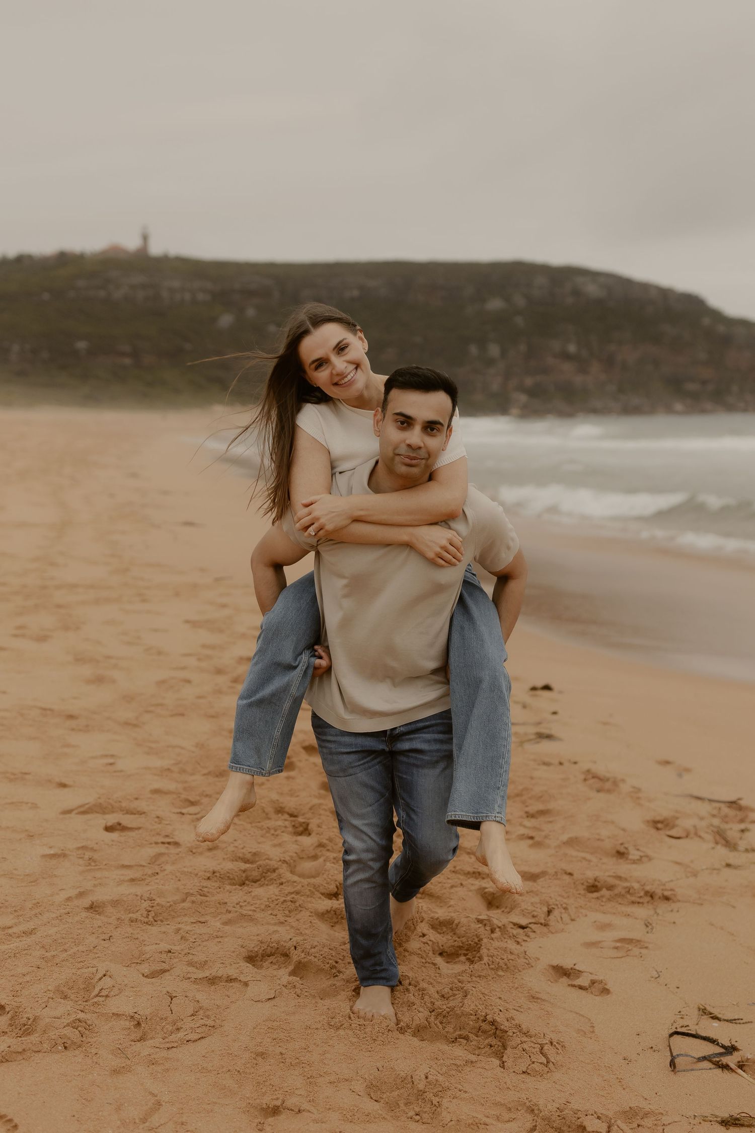 A couple plays and laughs together while running on a sandy beach during an overcast day.