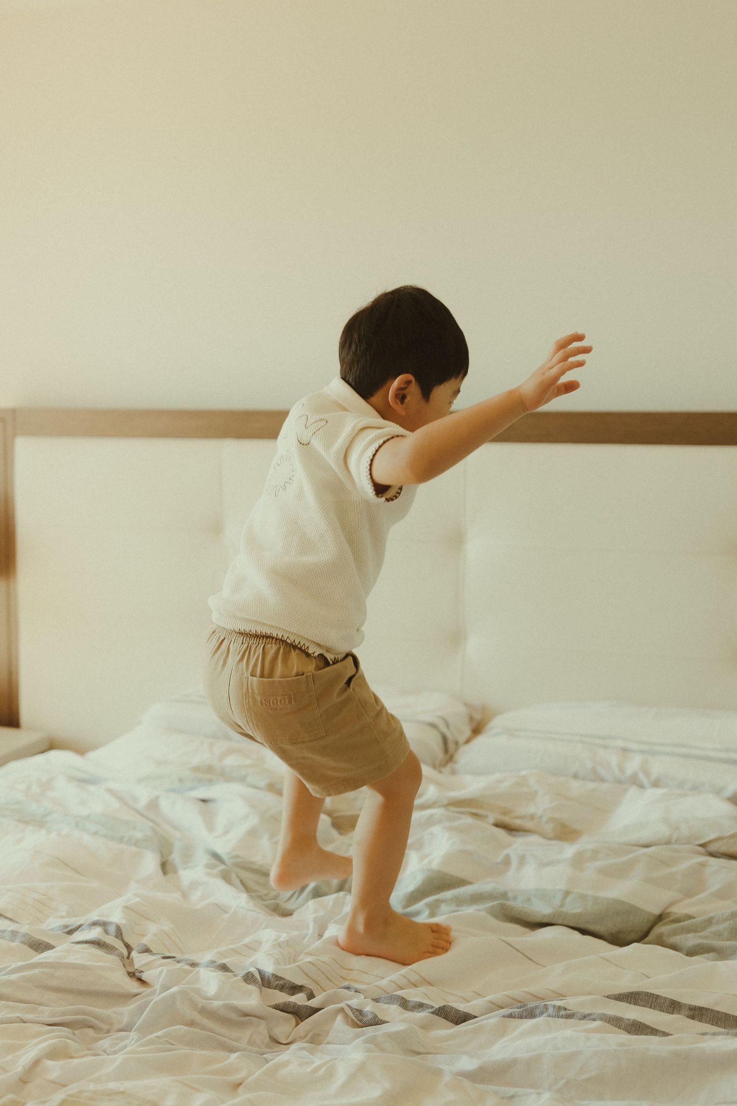A young child jumps playfully on a white bed with unmade sheets.