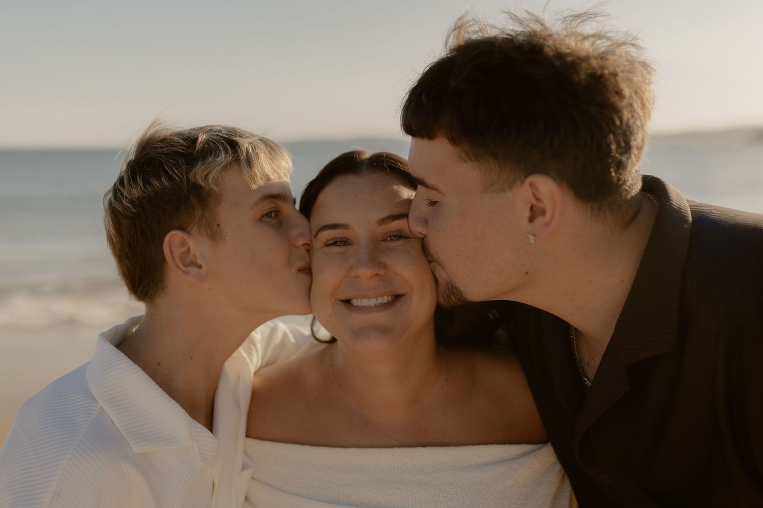 Three people share an intimate moment together against a sunlit coastal backdrop.