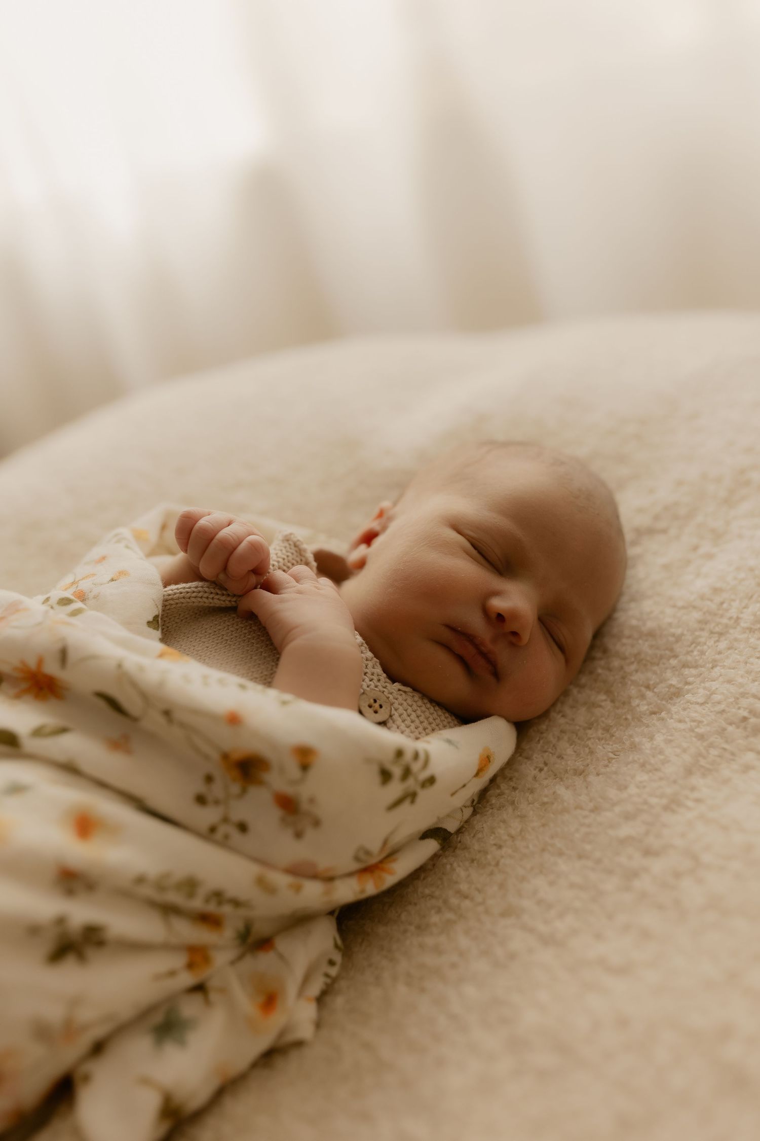 A sleeping newborn baby wearing a floral outfit rests peacefully on cream-colored bedding.