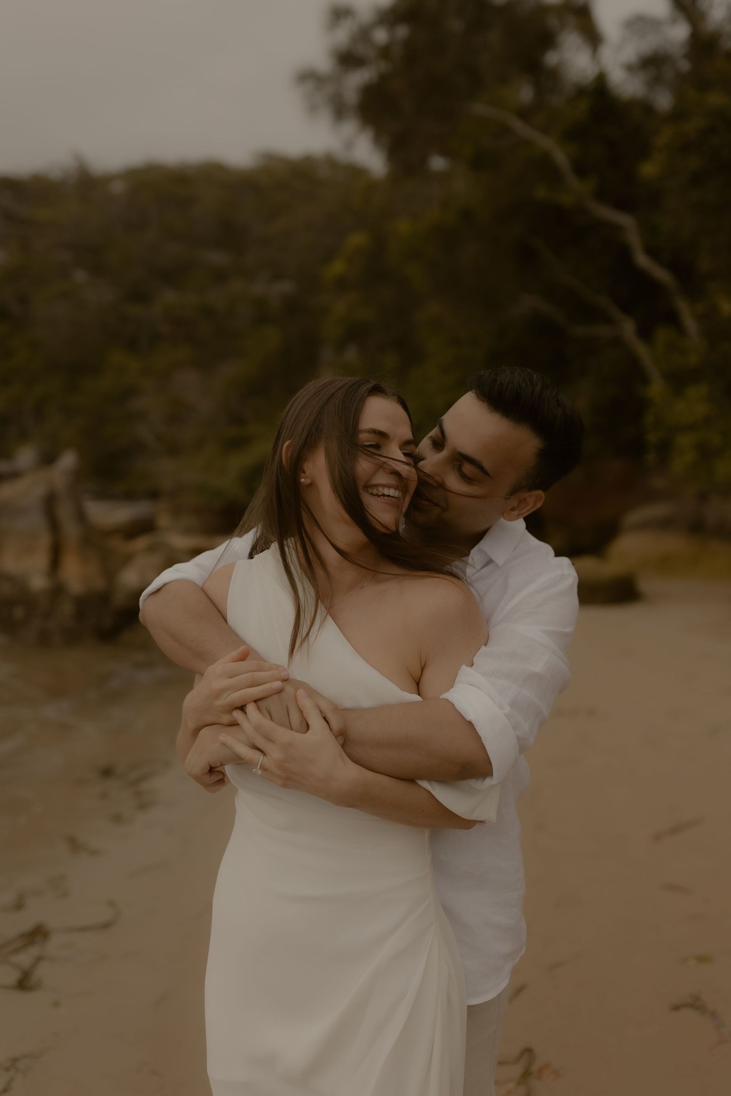 A romantic couple embraces on a beach during sunset, with the woman wearing a flowing white dress.