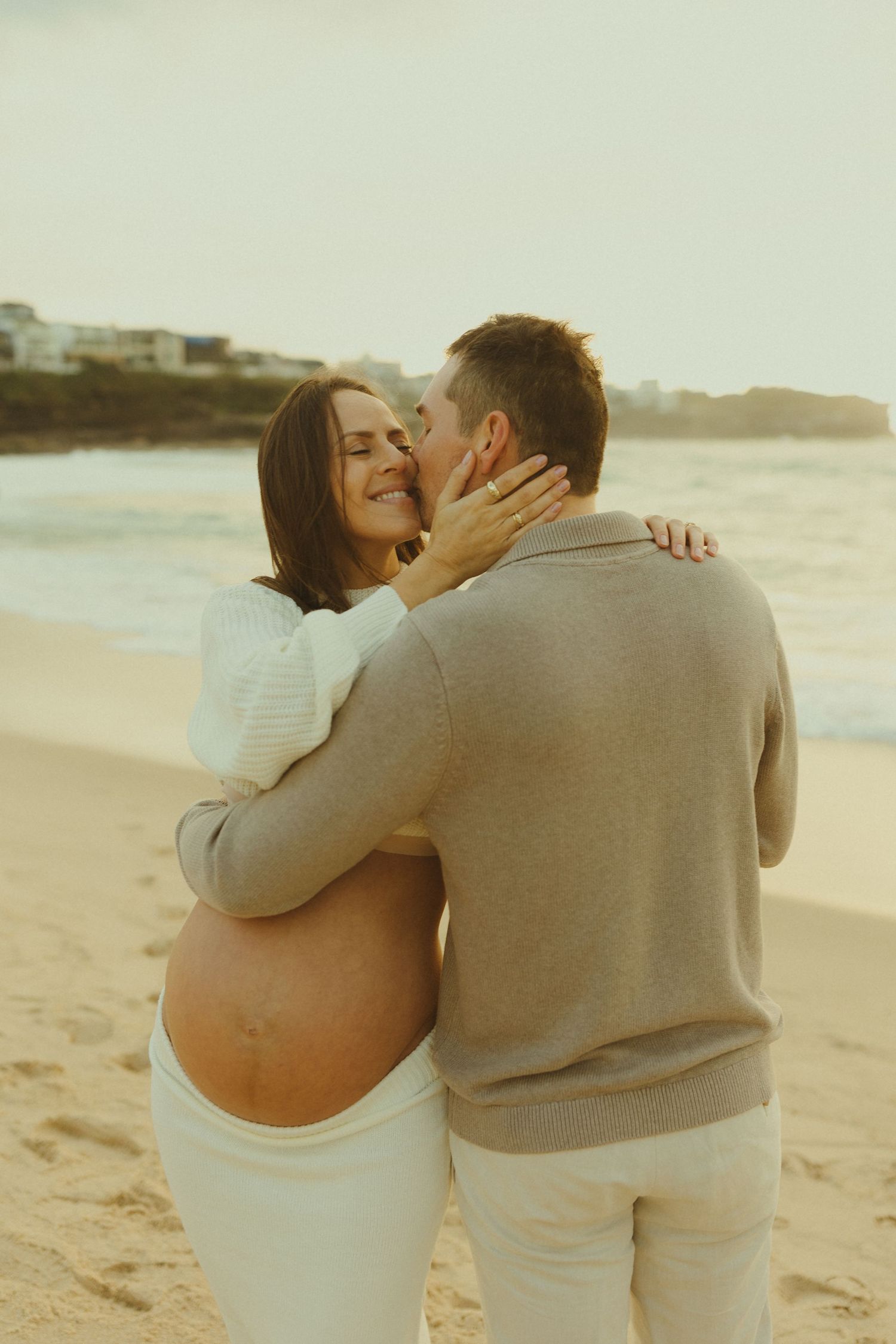 An expectant couple embracing tenderly on the beach with the pregnant woman wearing white and her partner in grey.
