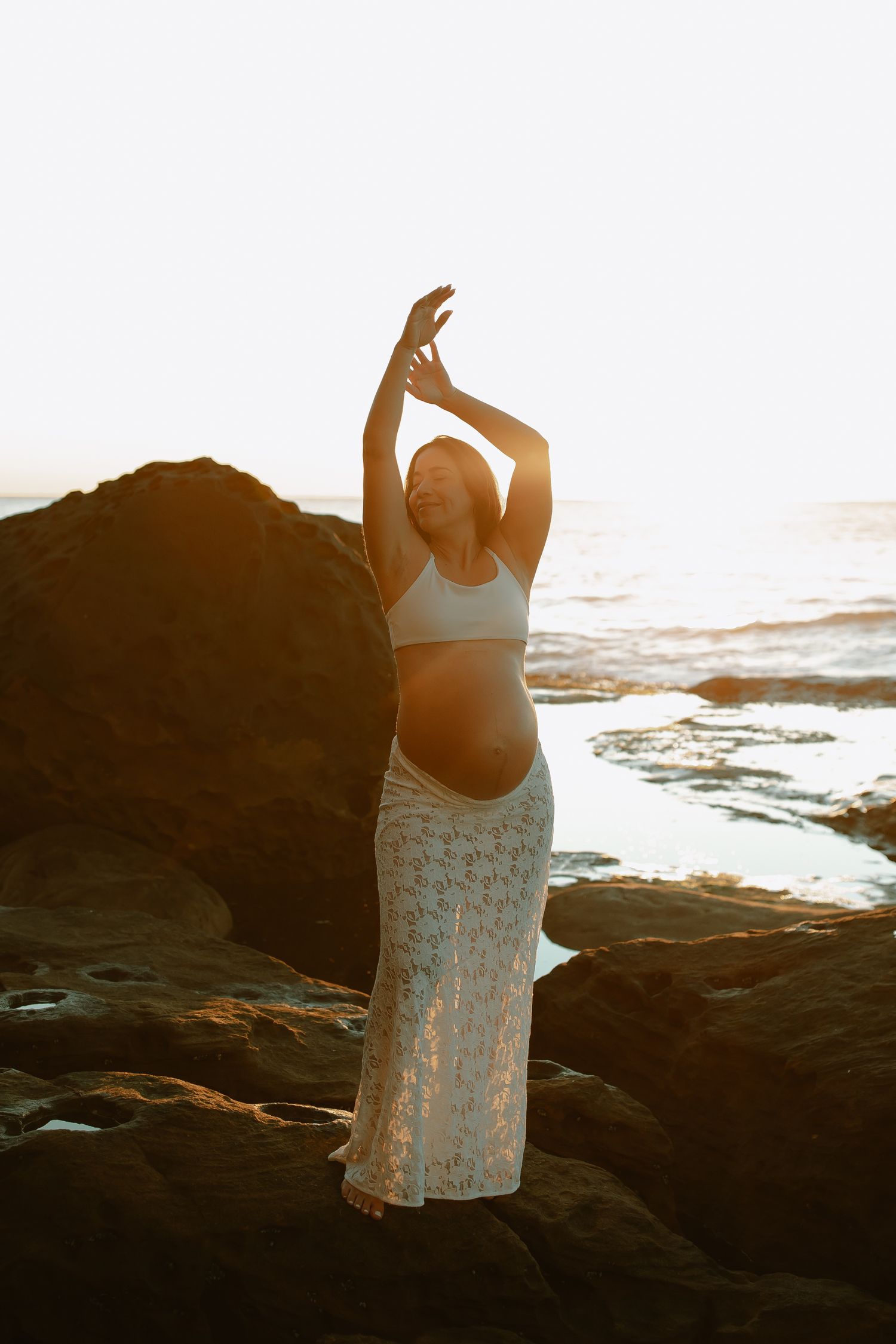 Silhouetted figure in a flowing white lace skirt stands gracefully on rocky beach at golden sunset with arms raised.