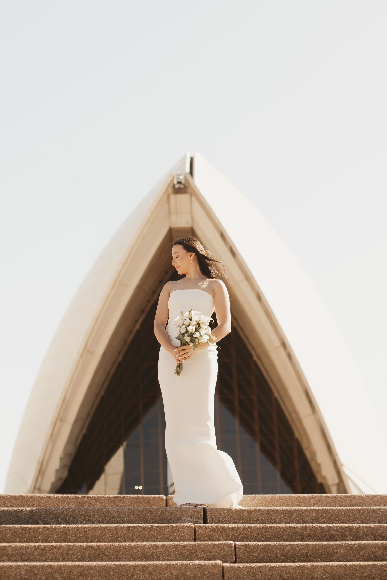 Bride holding white bouquet poses on steps of Sydney Opera House in fitted wedding gown.