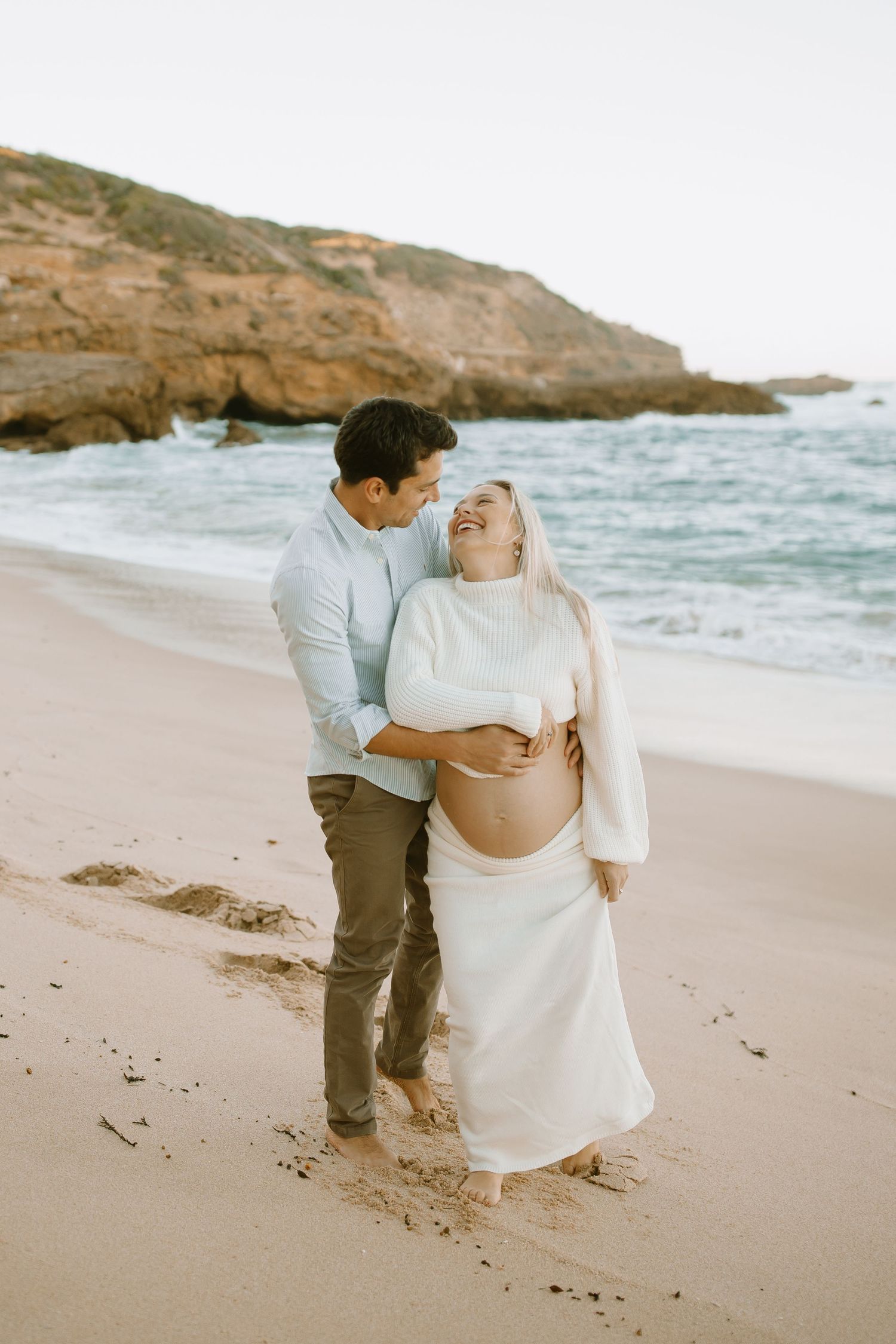 A couple embraces on a scenic beach with rocky cliffs as they pose for a maternity photoshoot.