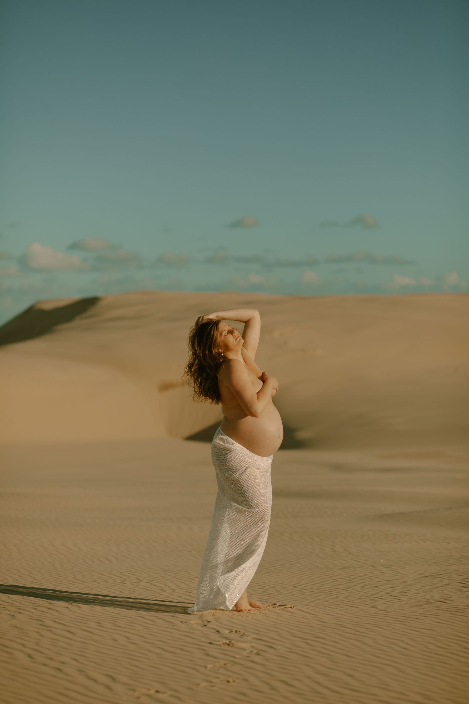 A dreamy desert maternity photoshoot features a woman in flowing white fabric standing on golden sand dunes at sunset.