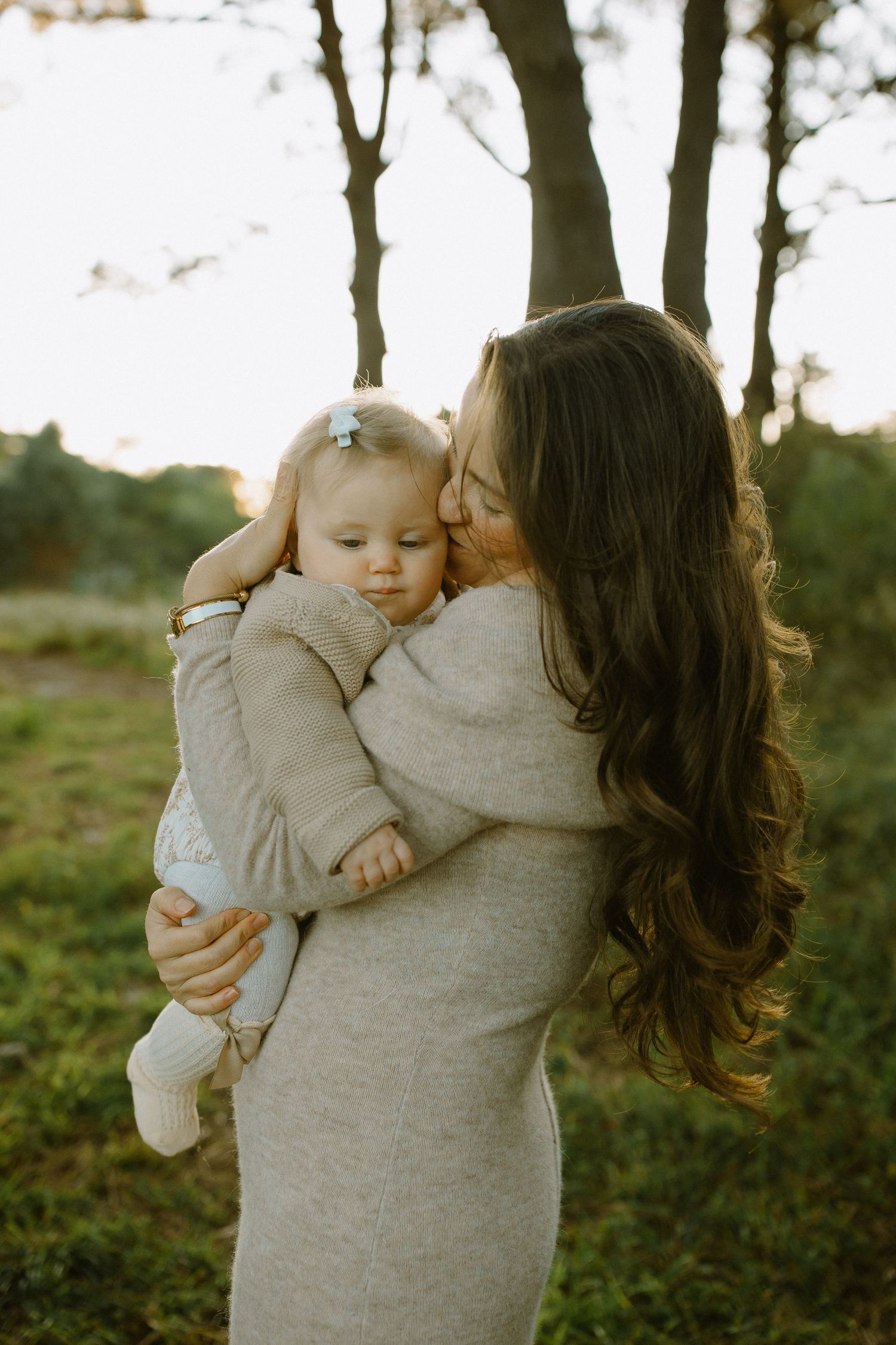 Woman in a beige sweater dress holds and cuddles a baby against her chest in a wooded setting.