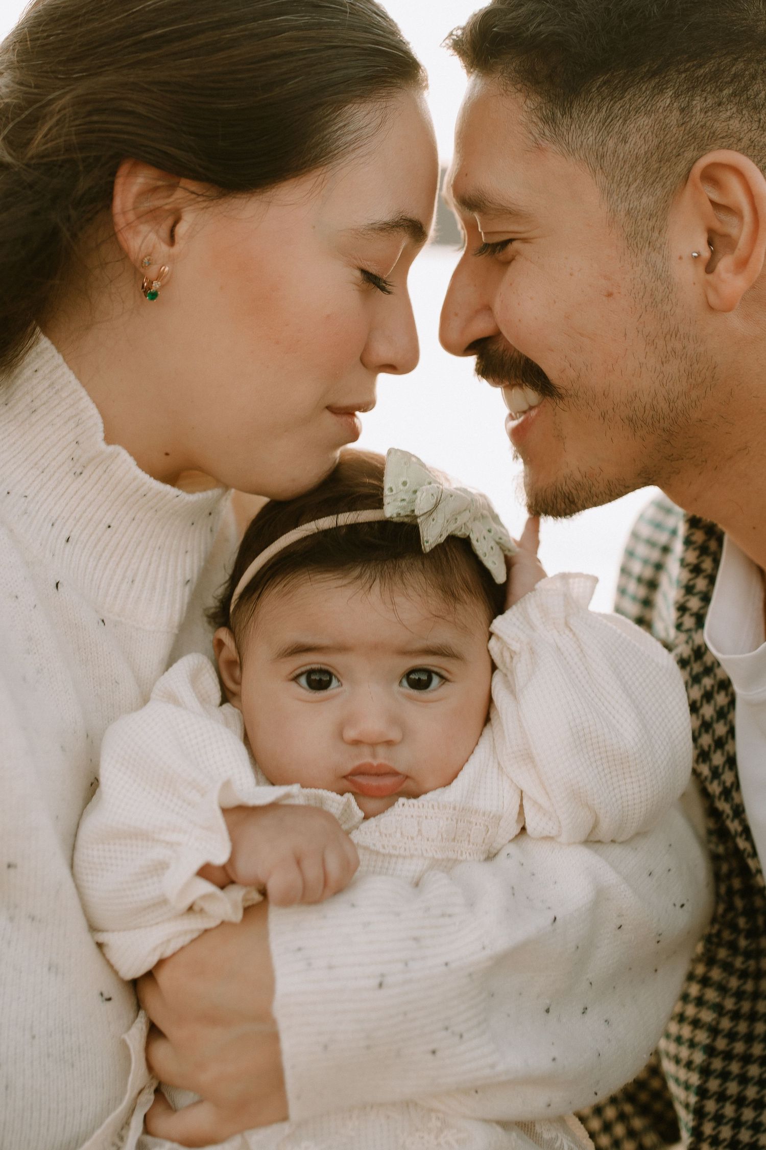 A warm family portrait showing parents lovingly embracing their baby wearing a white bow headband.