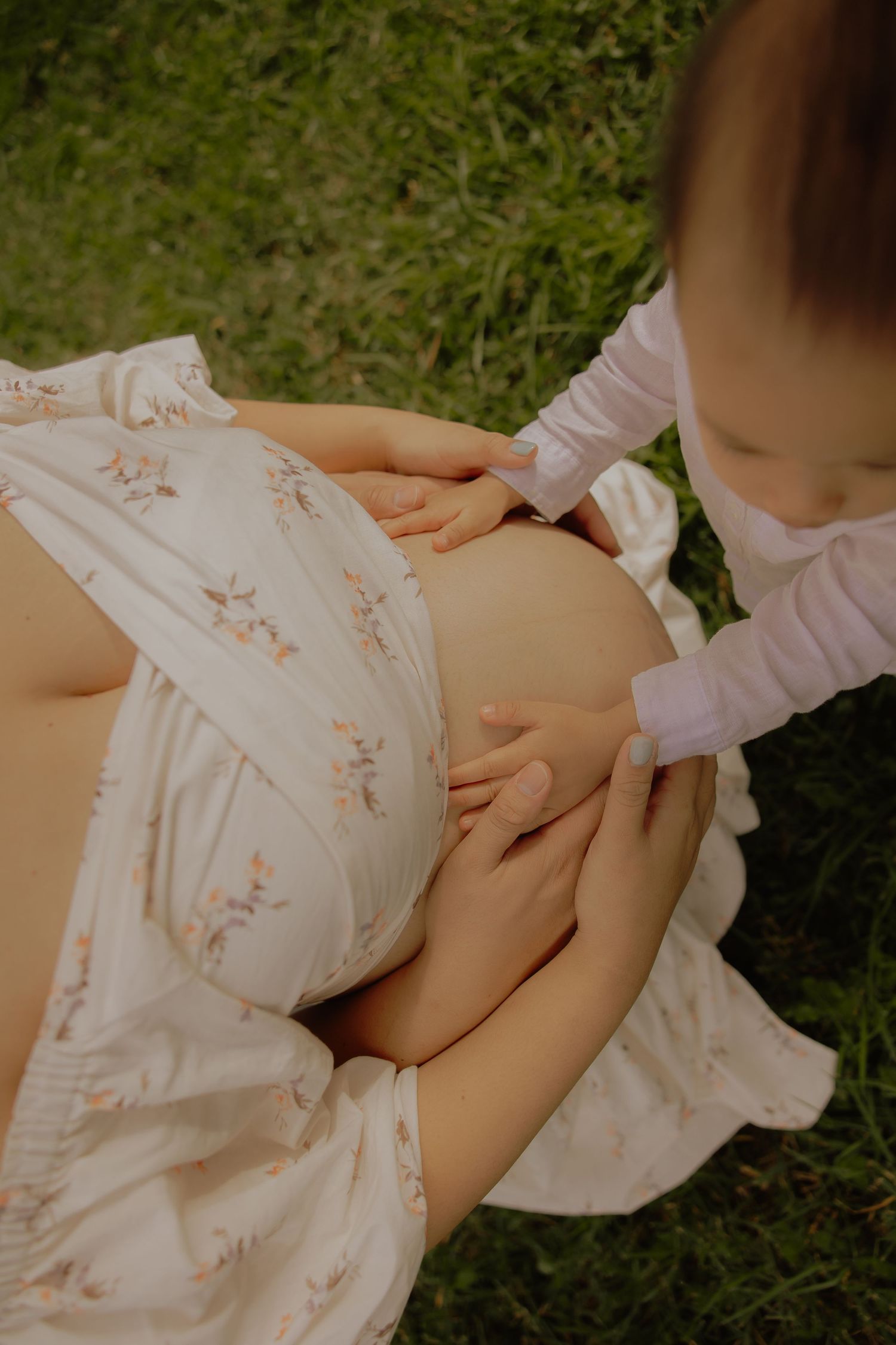 Close-up of toddler gently touching pregnant mothers bare baby bump while sitting on grass outdoors.