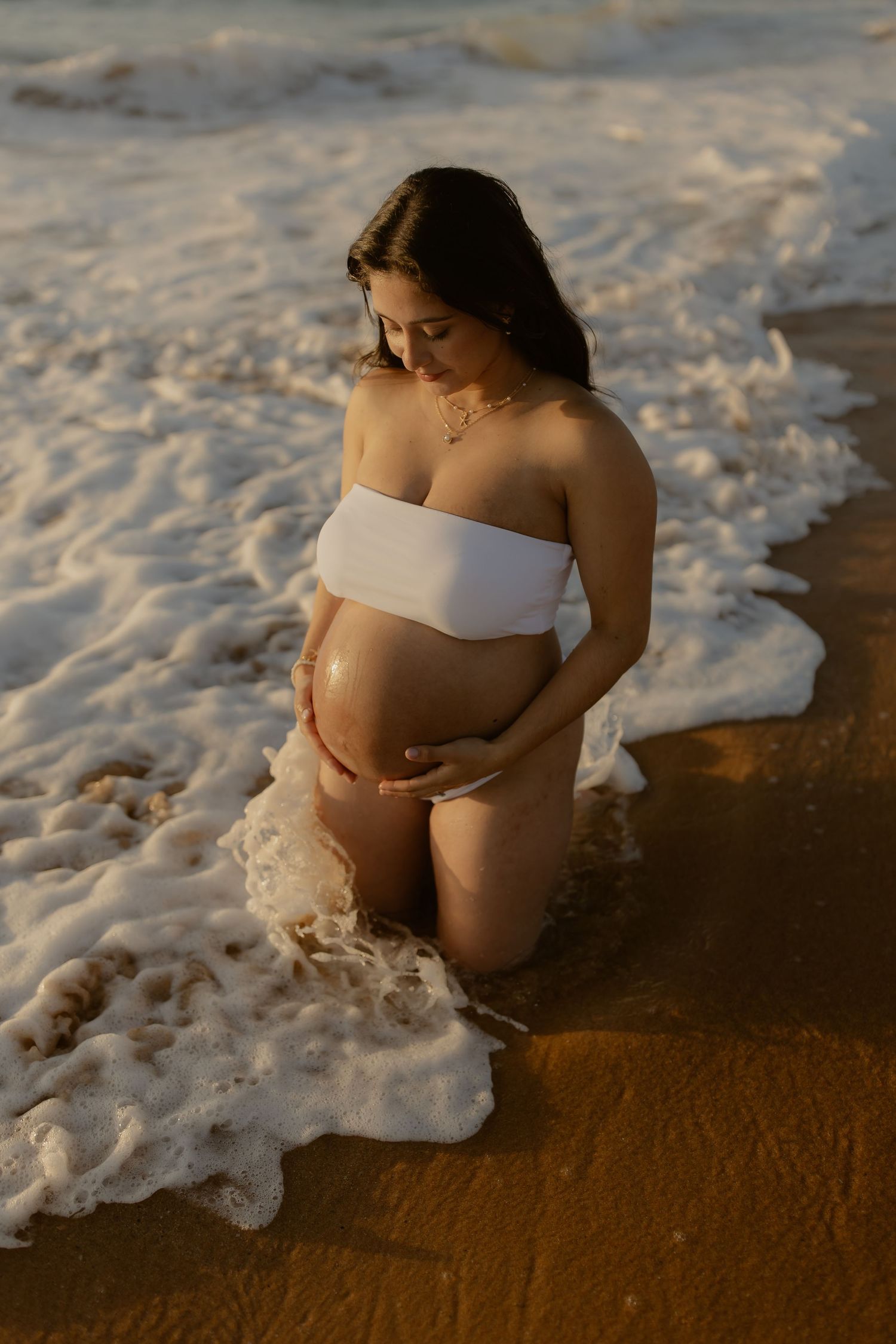 A pregnant woman in white swimwear sits on a sandy beach as ocean waves gently roll in during golden hour.