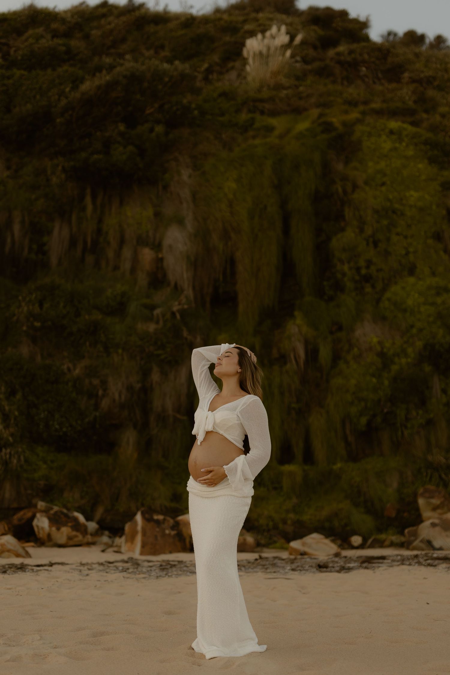 A pregnant woman in a white dress poses on a beach with dramatic cliffs in the background during golden hour.