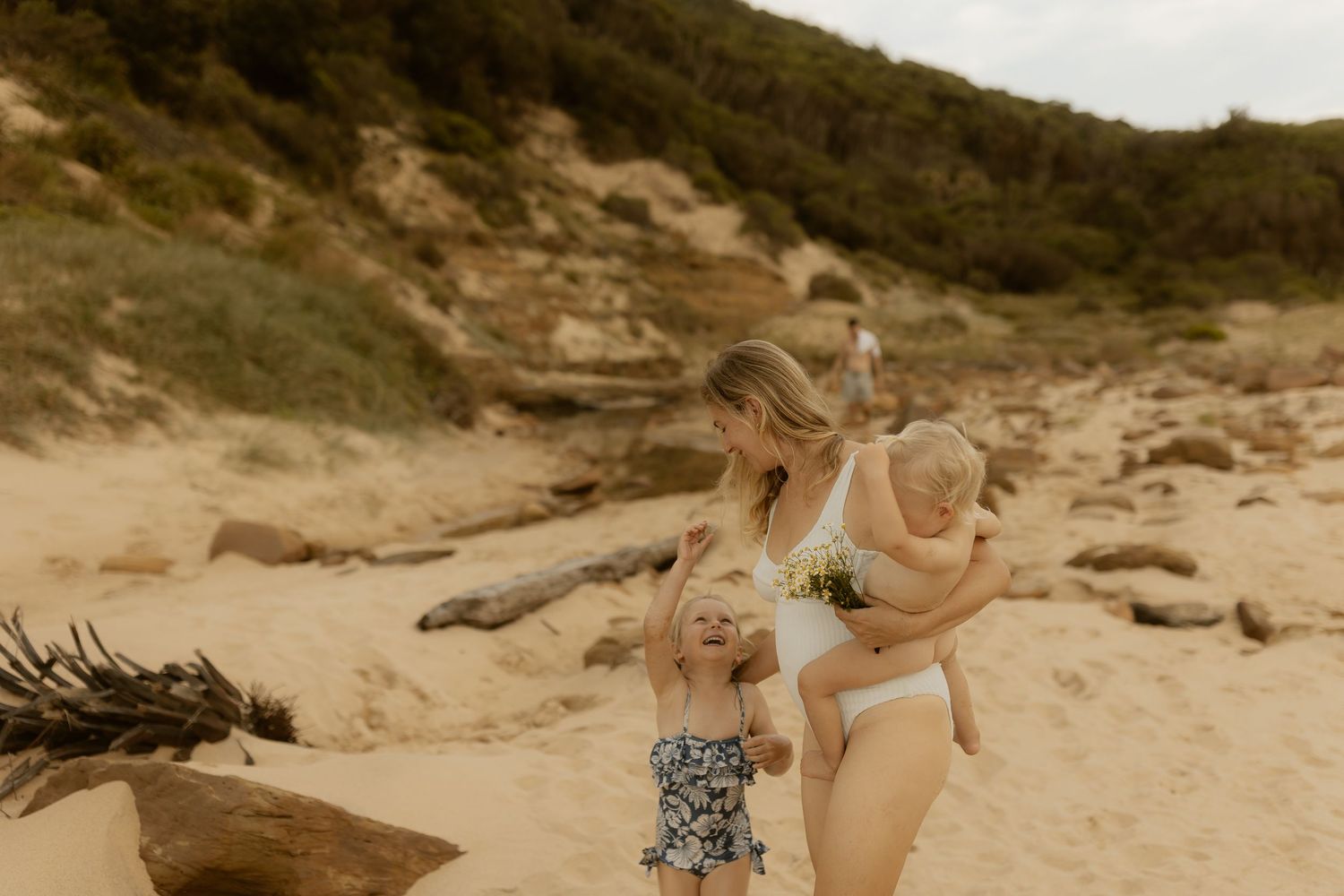 A sunny beach scene with figures enjoying time together among sand dunes and driftwood.