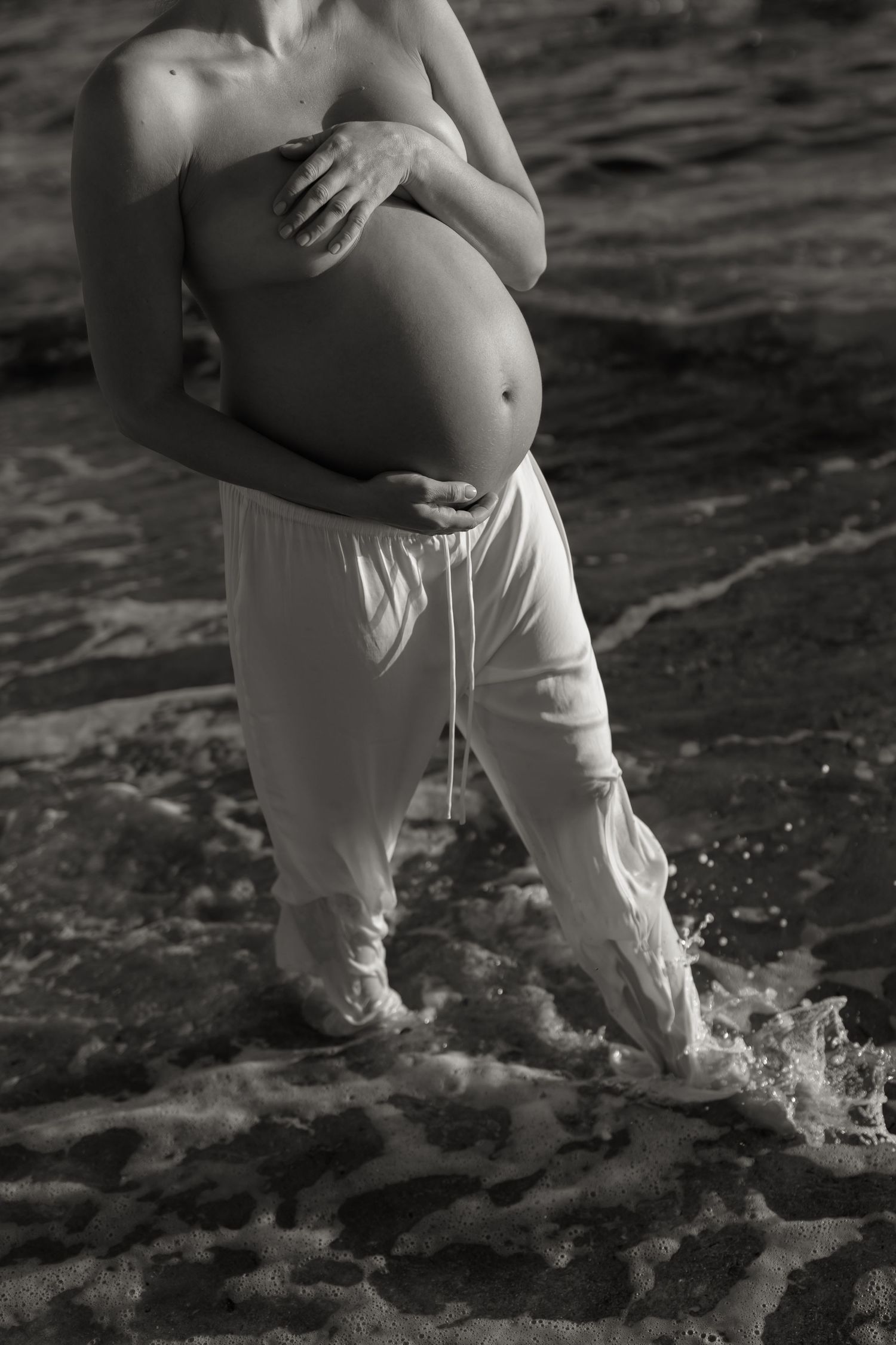 Black and white artistic maternity photo of a pregnant belly with waves splashing at the feet.