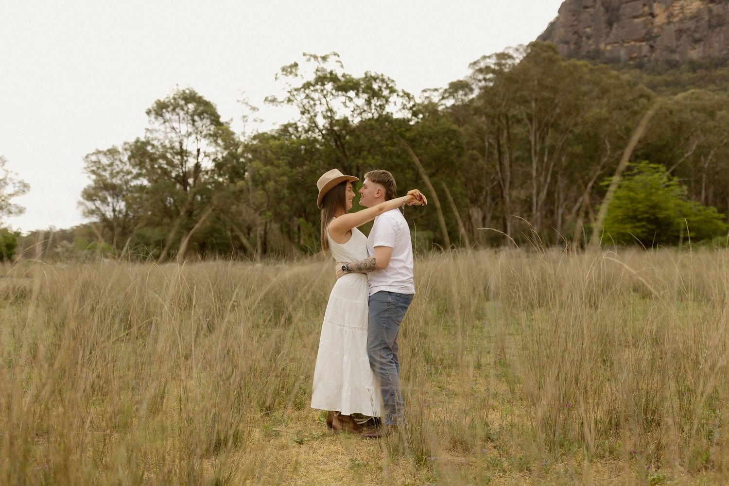 A couple embraces in a golden field with mountains and eucalyptus trees in the background at sunset.