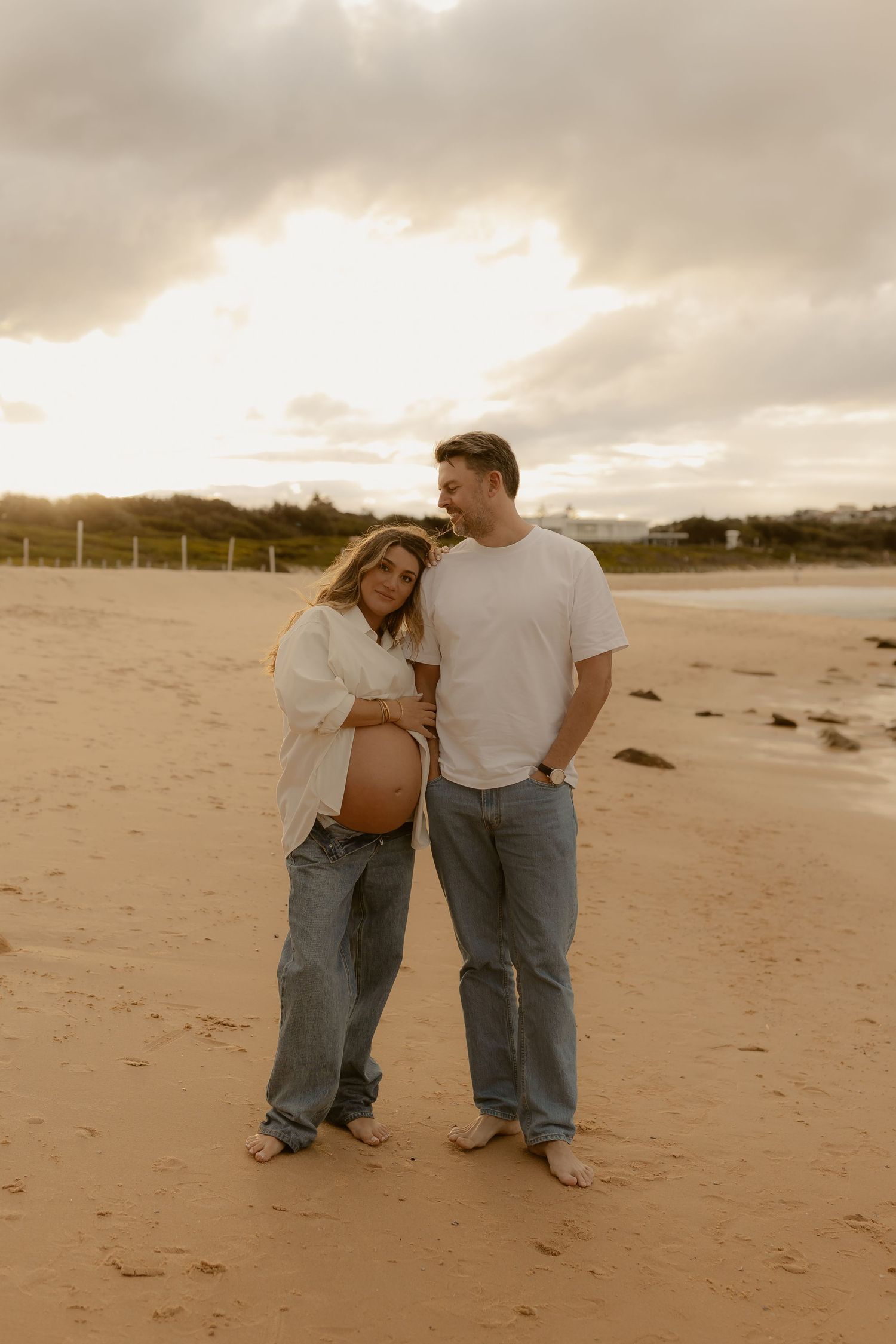 Couple maternity photo on sandy beach at sunset, pregnant woman in white shirt and jeans with partner.