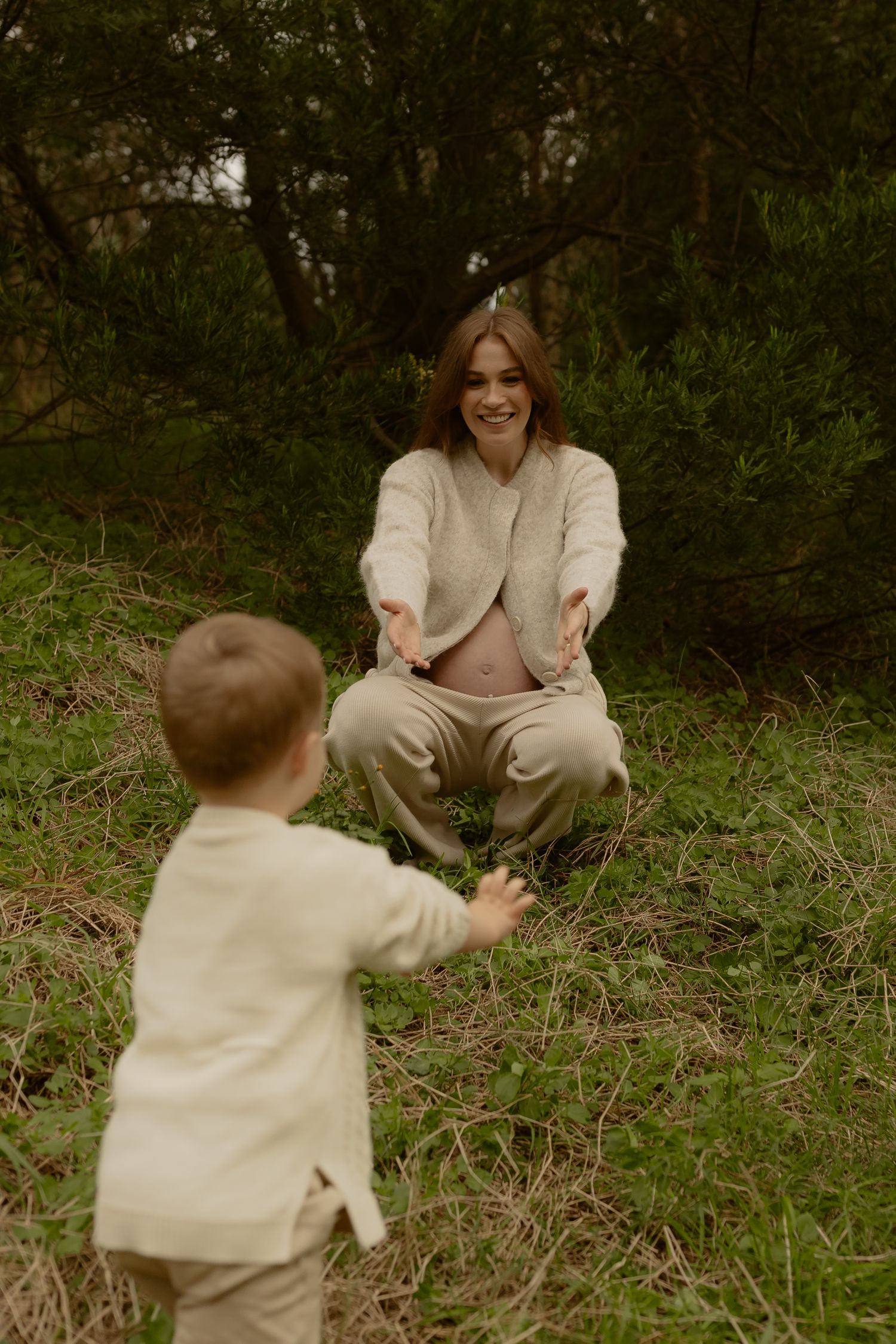 Smiling pregnant woman crouches with open arms as her toddler runs toward her in a forest clearing.